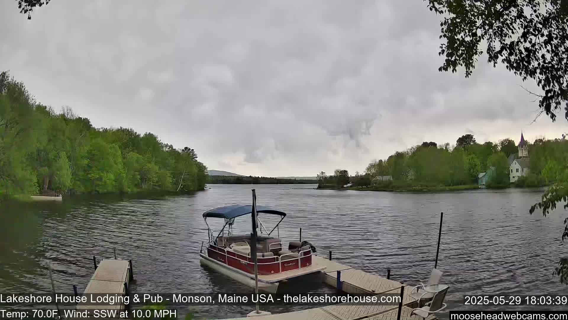 A pontoon boat is docked at a lakeside pier under an overcast sky, with a calm lake surrounded by lush green trees and distant buildings.