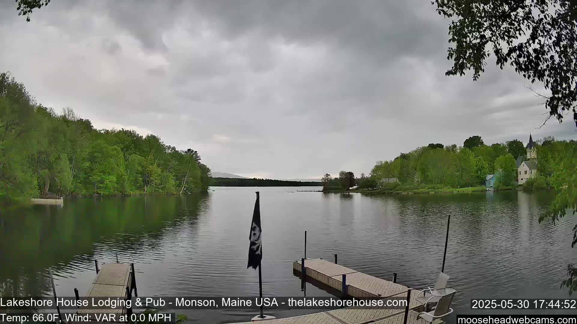 A calm lake under an overcast sky is viewed from a wooden dock, with lush green trees lining the shore and buildings visible in the distance.
