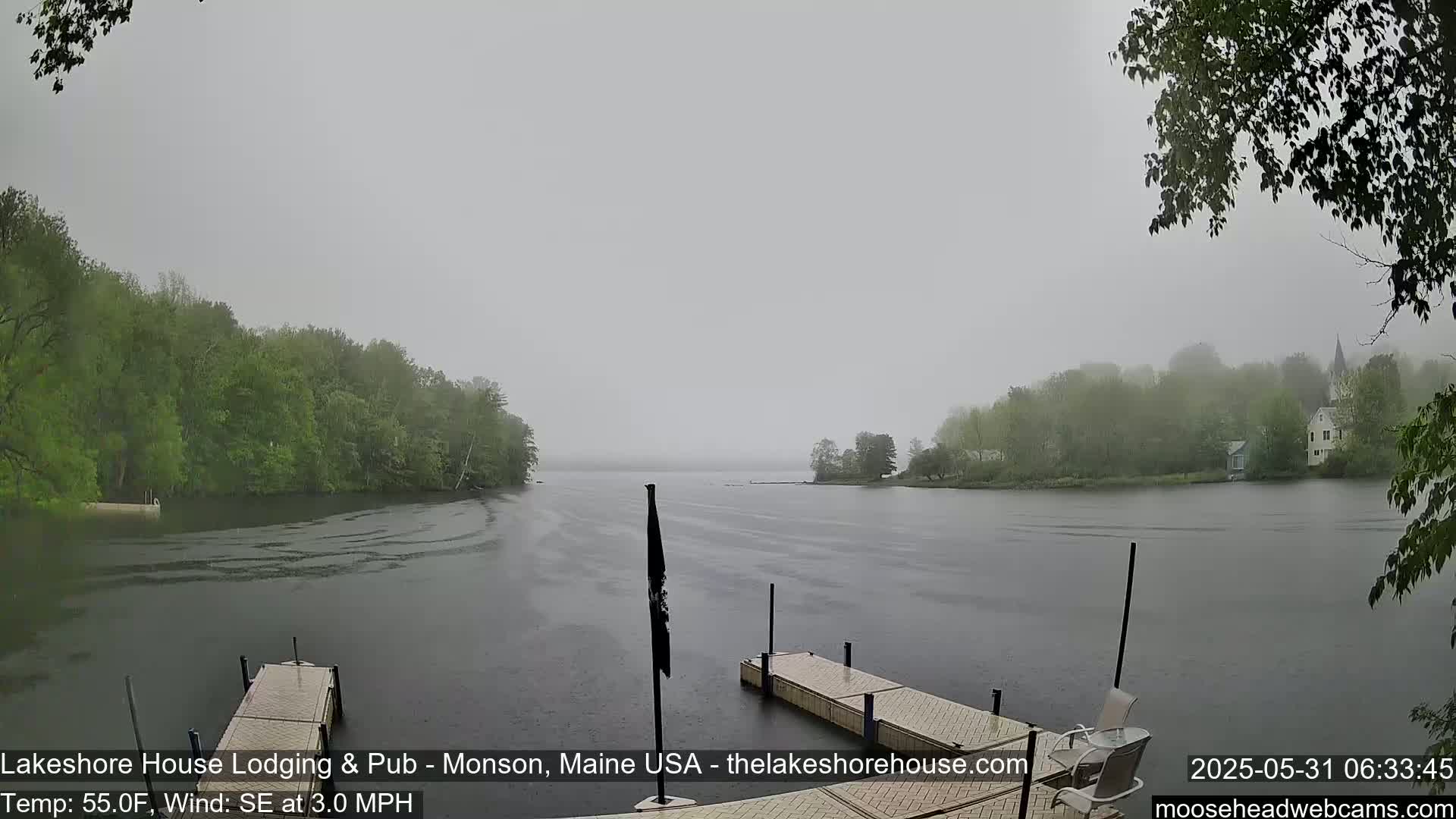 A calm lake, shrouded in a misty rain, is seen from a lakeside dock, with green trees lining the shore.