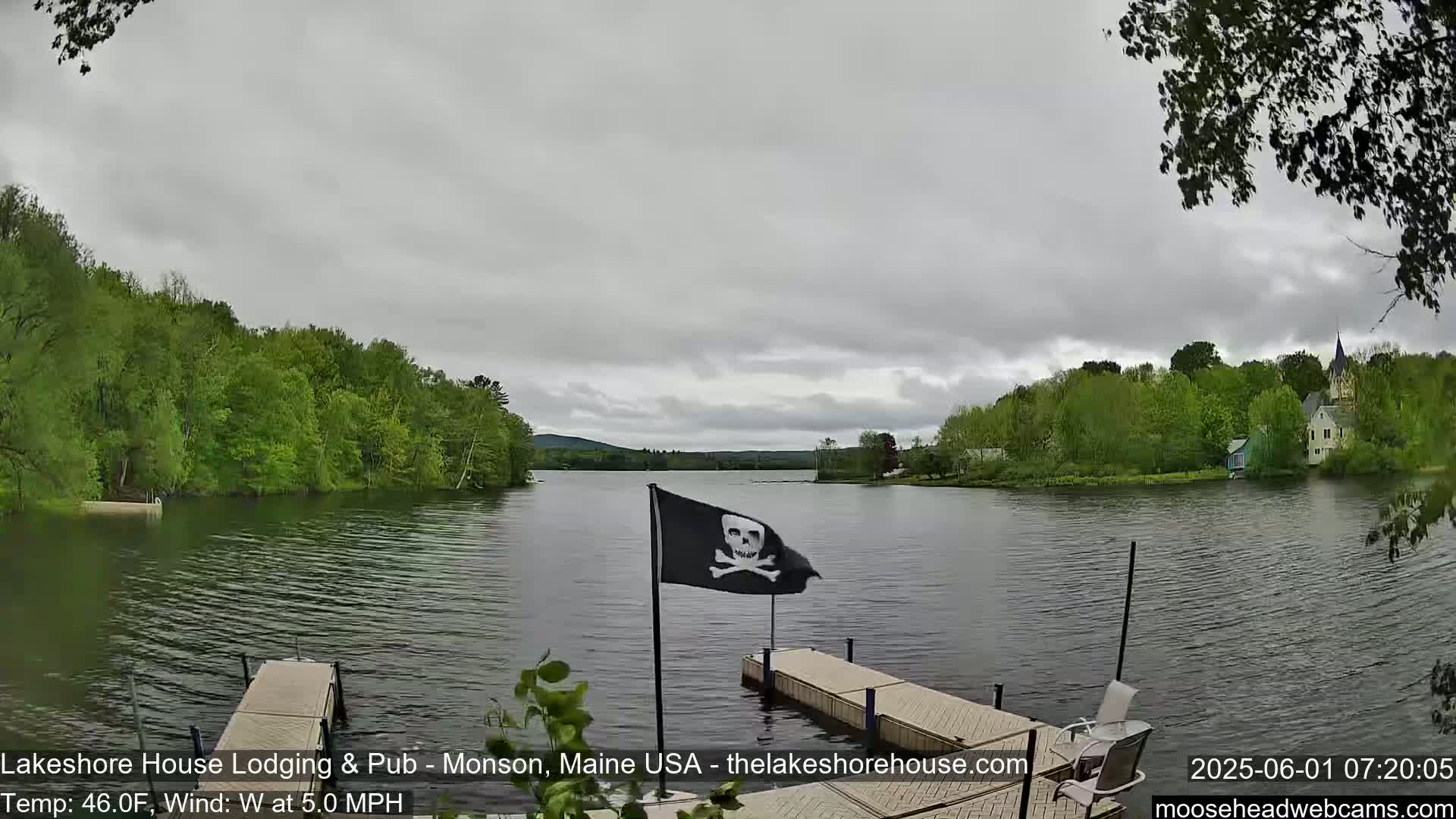 A calm lake with green, tree-lined shores, a pirate flag on a dock, and an overcast sky.