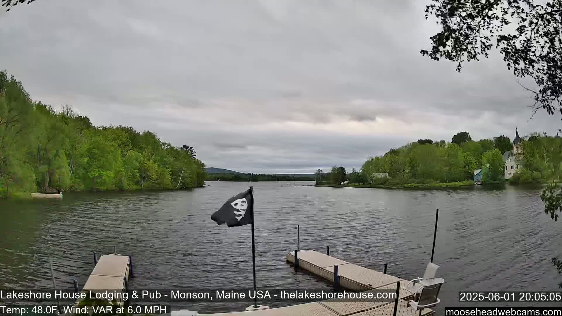 A calm lake, under an overcast sky, is viewed from a dock with a pirate flag flying, and trees and buildings line the shore in the background.
