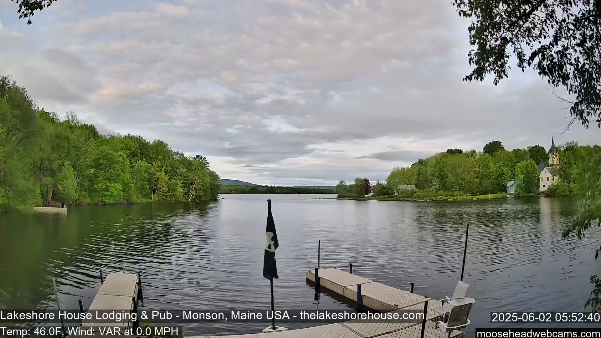A calm lake is seen from a wooden dock, with lush green trees lining the shores and a cloudy sky overhead.