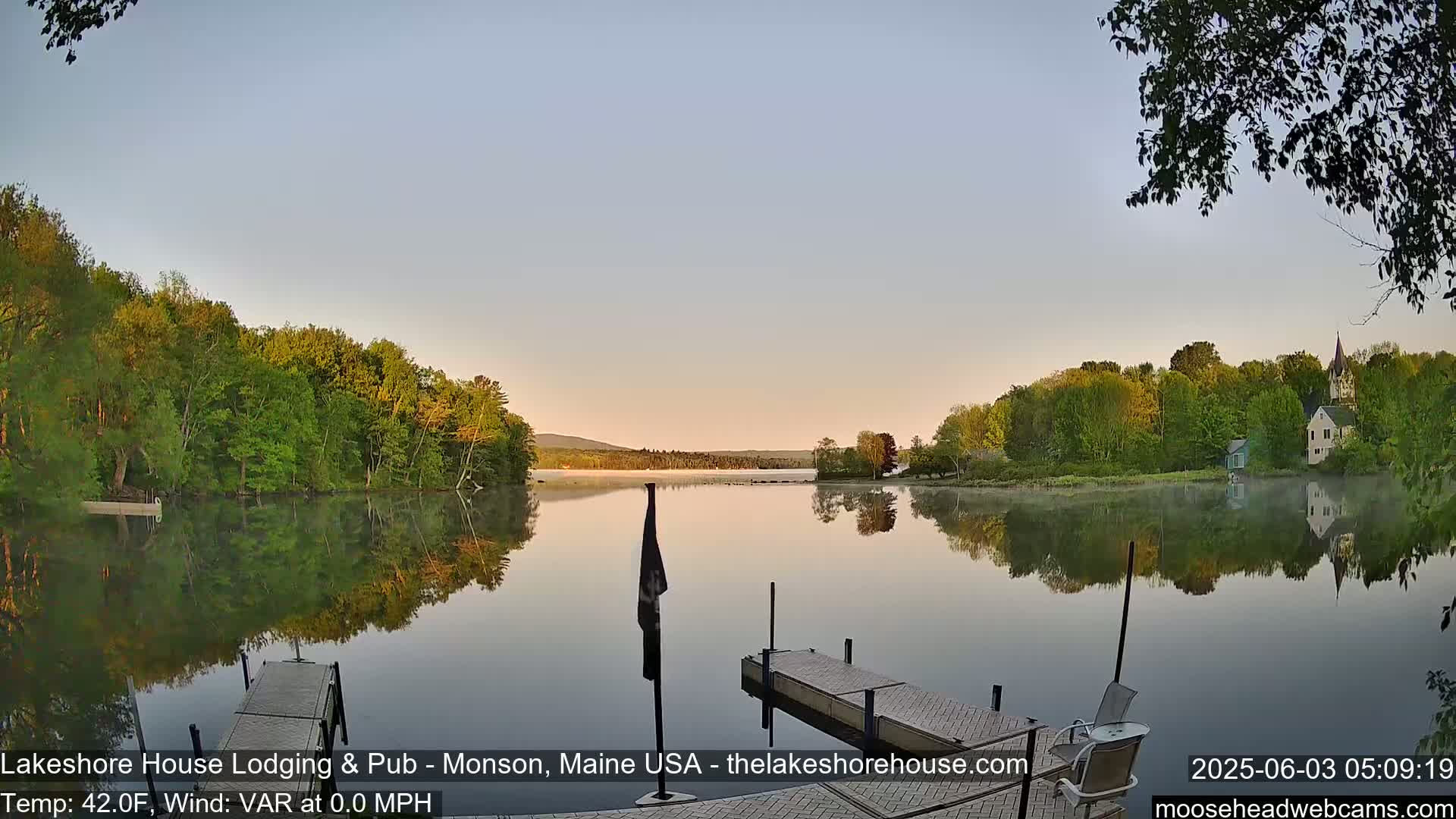 A calm lake reflects the trees and buildings on its shore at sunrise, with a wooden dock in the foreground.