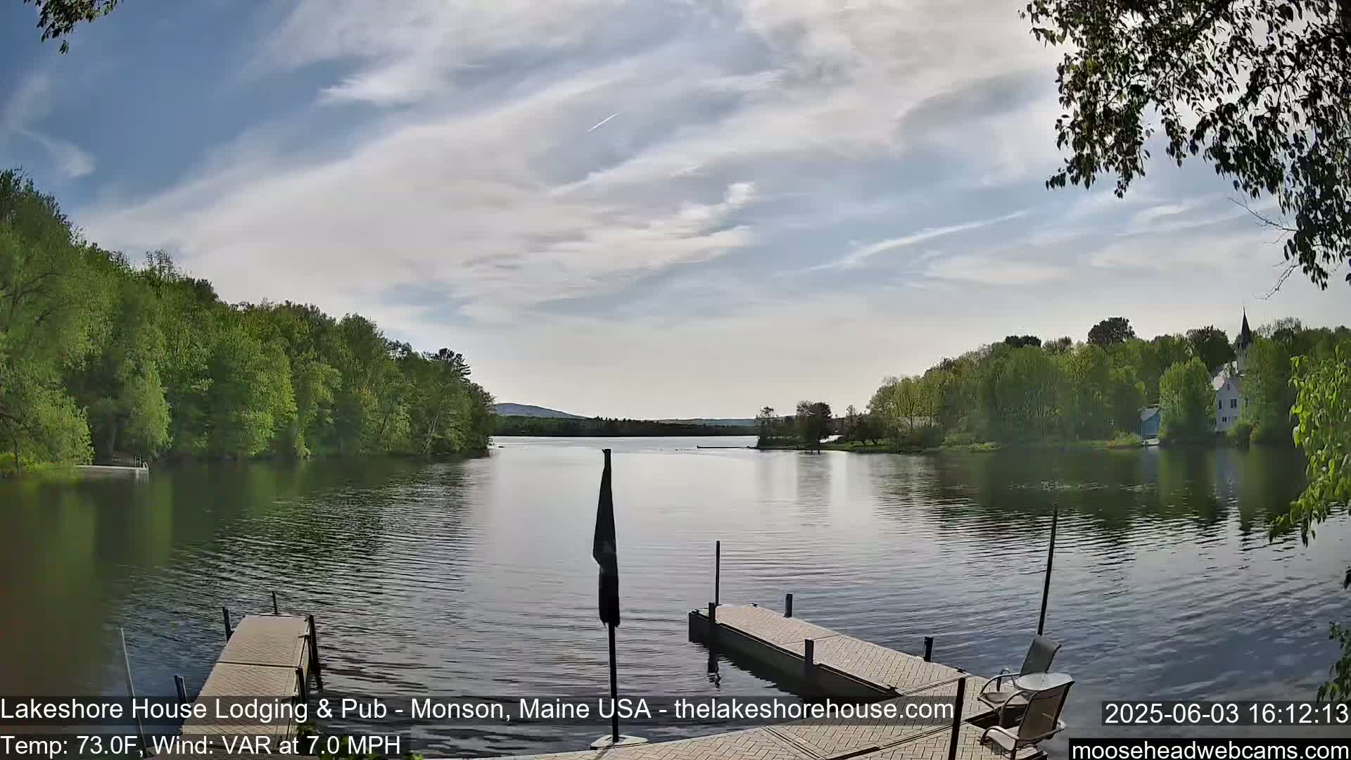 A calm lake with a wooden dock is seen, surrounded by lush green trees under a partly cloudy sky.