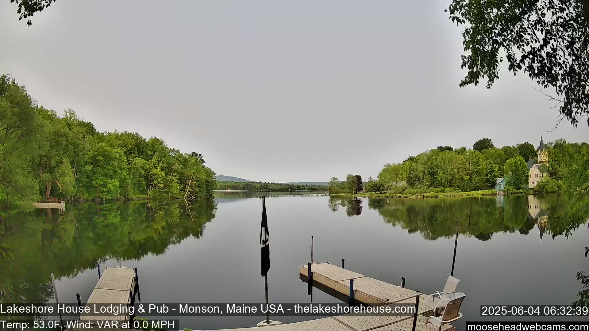 A calm lake reflects the green trees and buildings on its shores under an overcast sky.