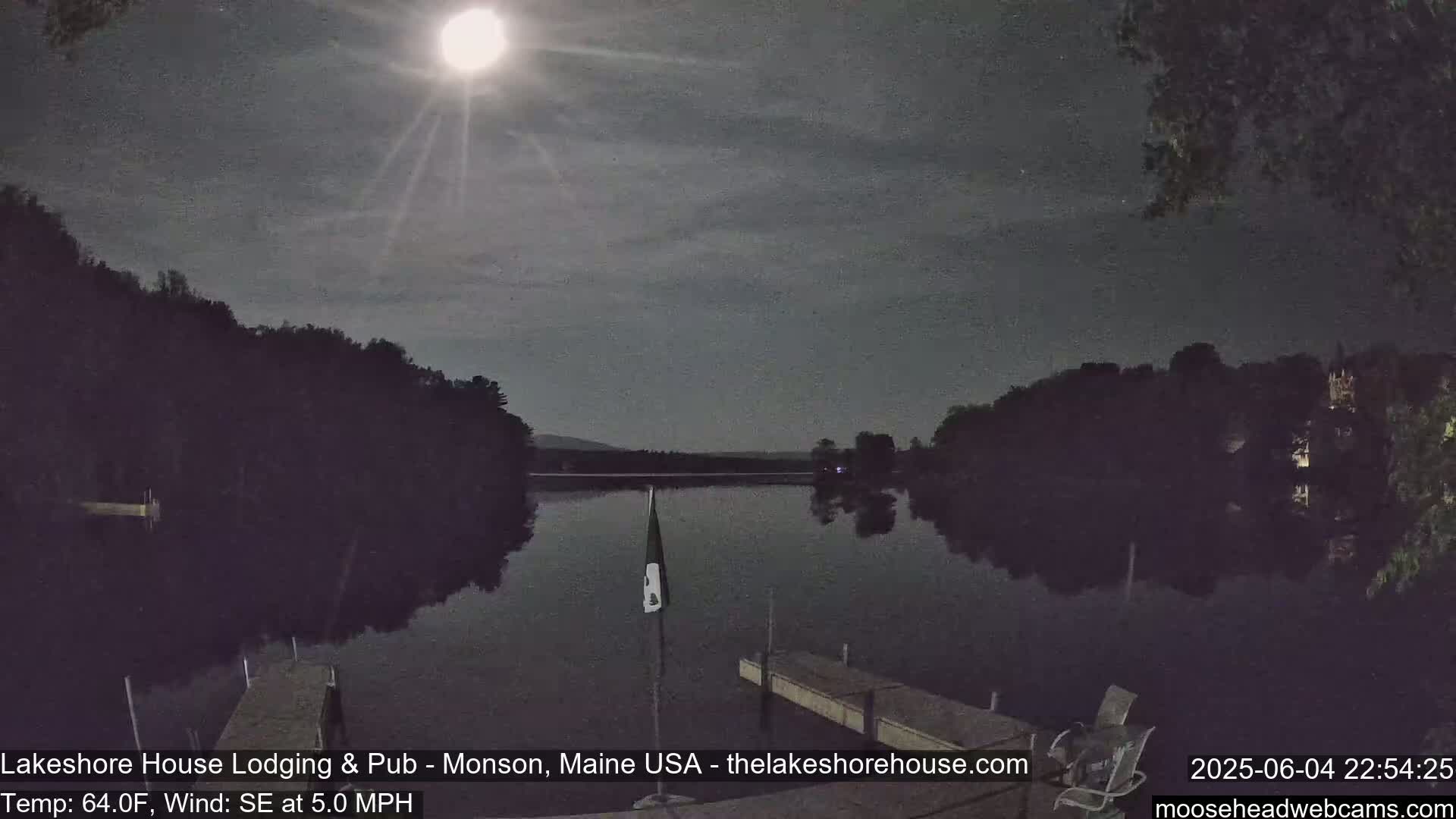 A bright moon illuminates a calm lake at night, surrounded by dark trees and a dock.