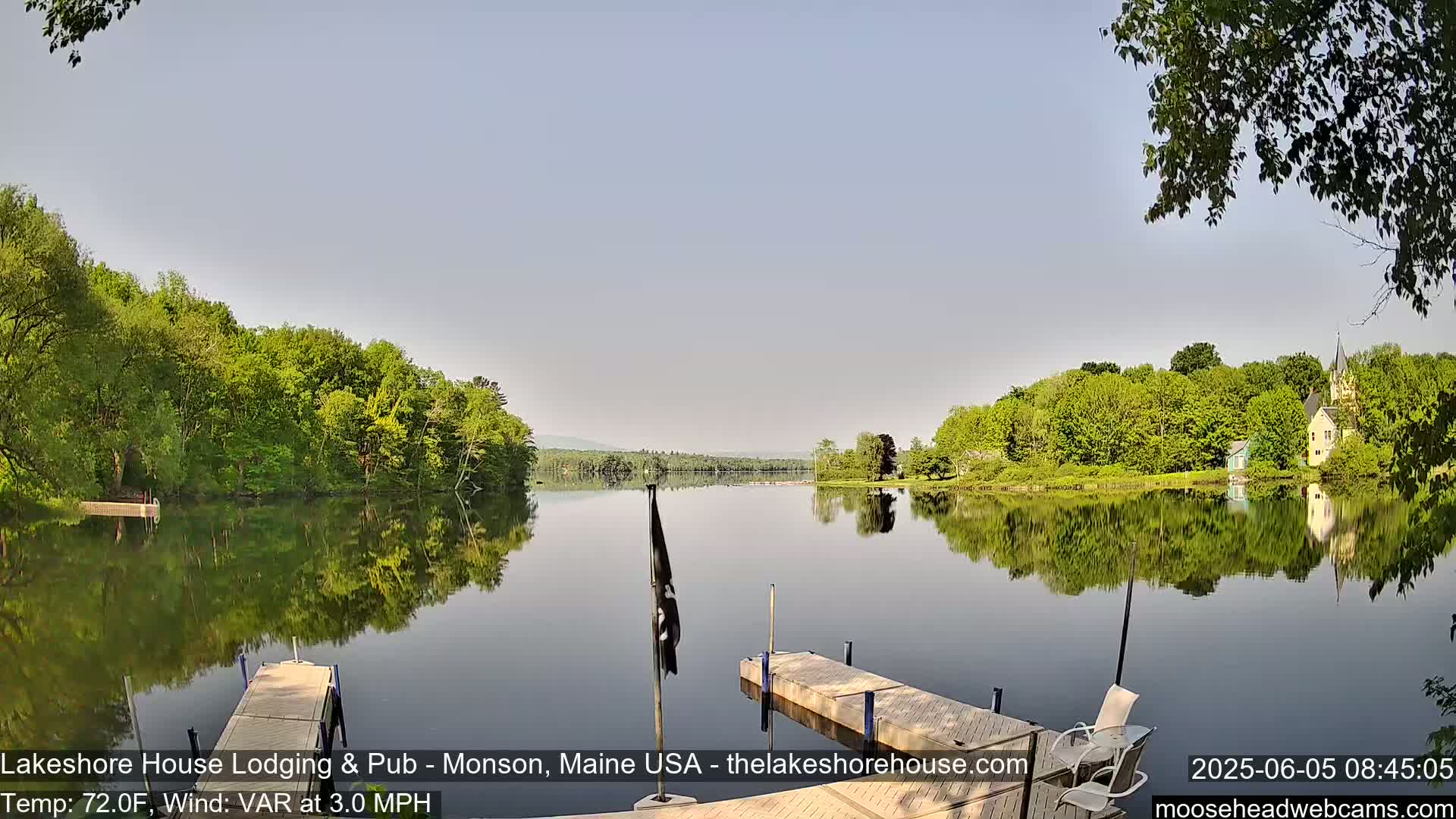 A calm lake reflects a tree-lined shore and buildings under a clear, light-blue sky.