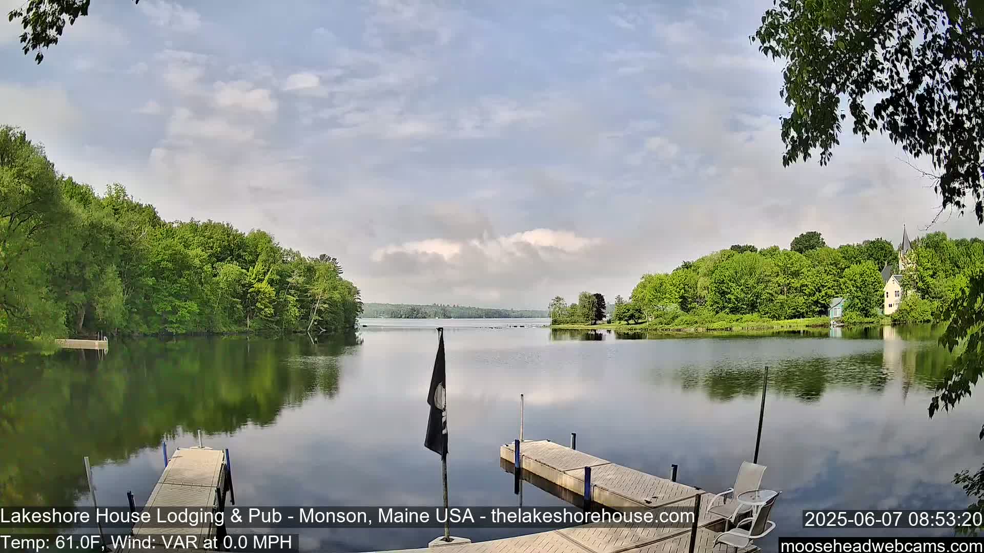 A calm lake with green trees lining its shores, a small dock in the foreground, and a partly cloudy sky.