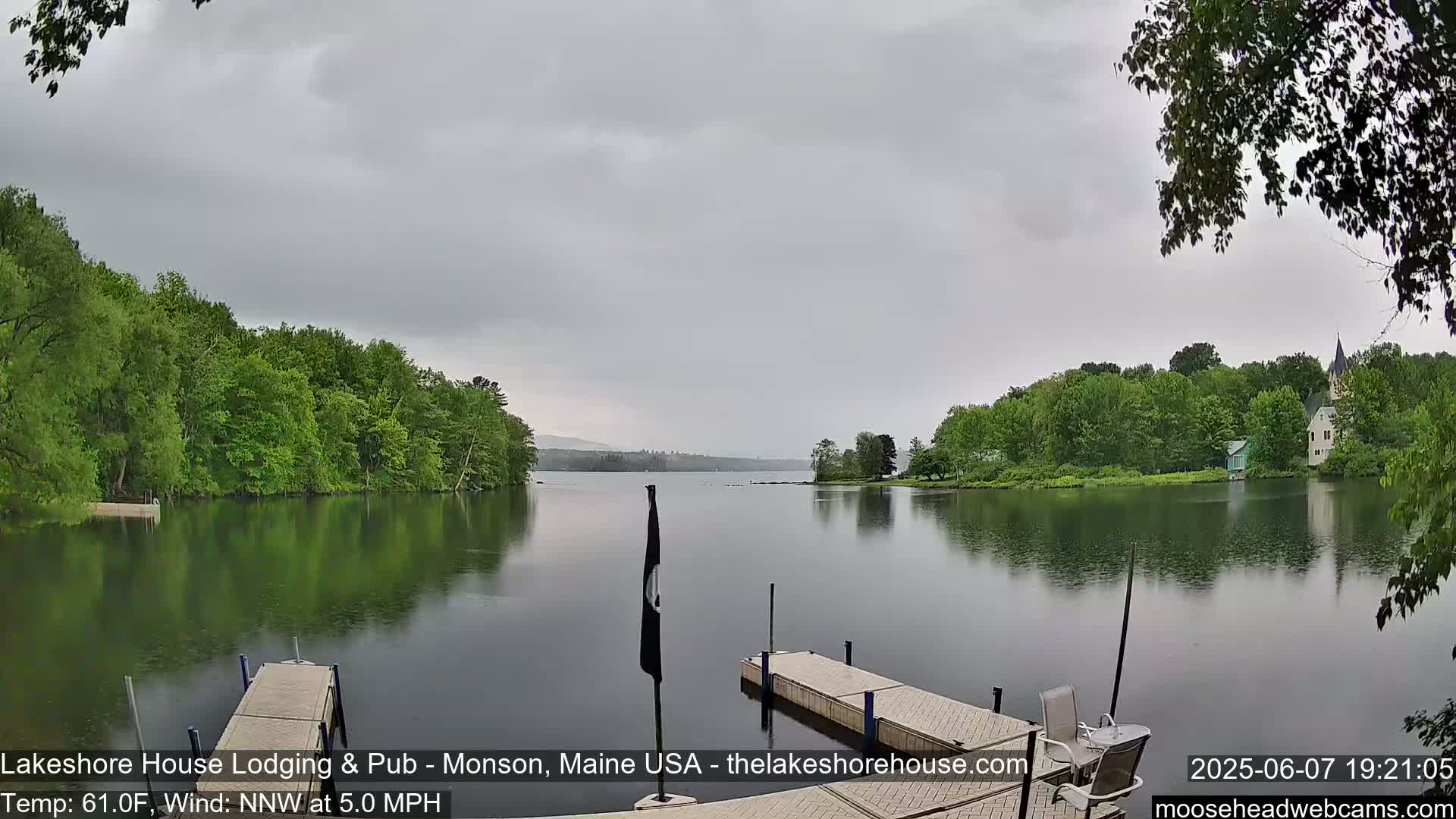 A calm lake under an overcast sky is bordered by lush green trees and features a dock with a small boat.