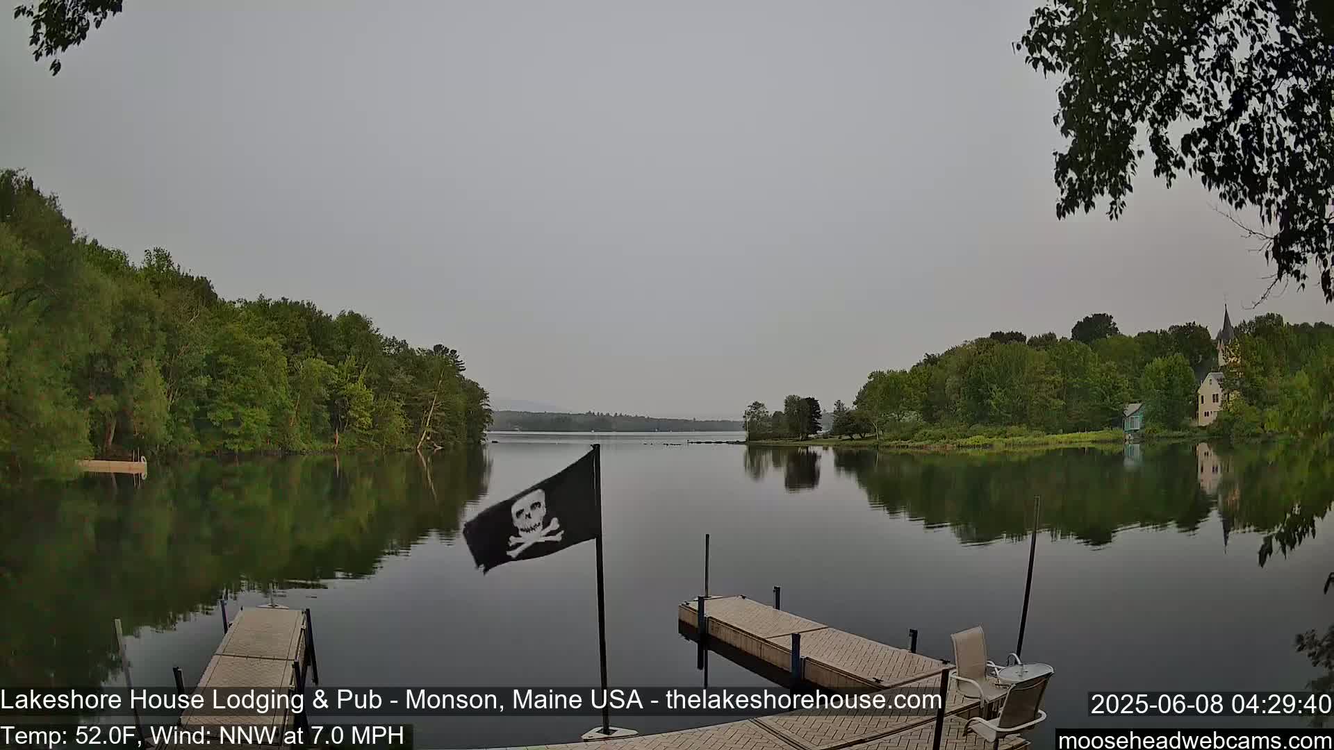 A calm lake reflects a gray sky and tree-lined shores, with a pirate flag and a wooden dock in the foreground.