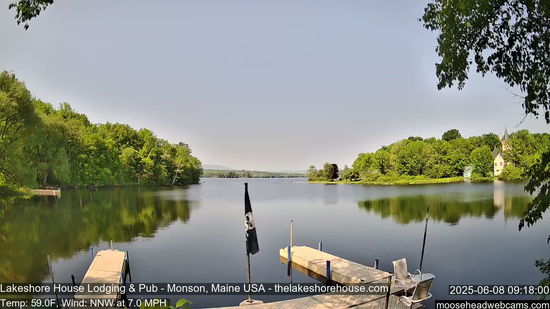A calm lake reflects the surrounding green trees and a distant building under a clear, light-blue sky.