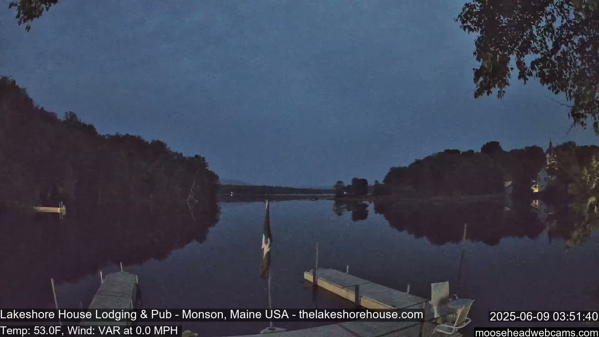 A calm, dark blue lake at night is reflected in the still water, bordered by dark trees and a small dock.