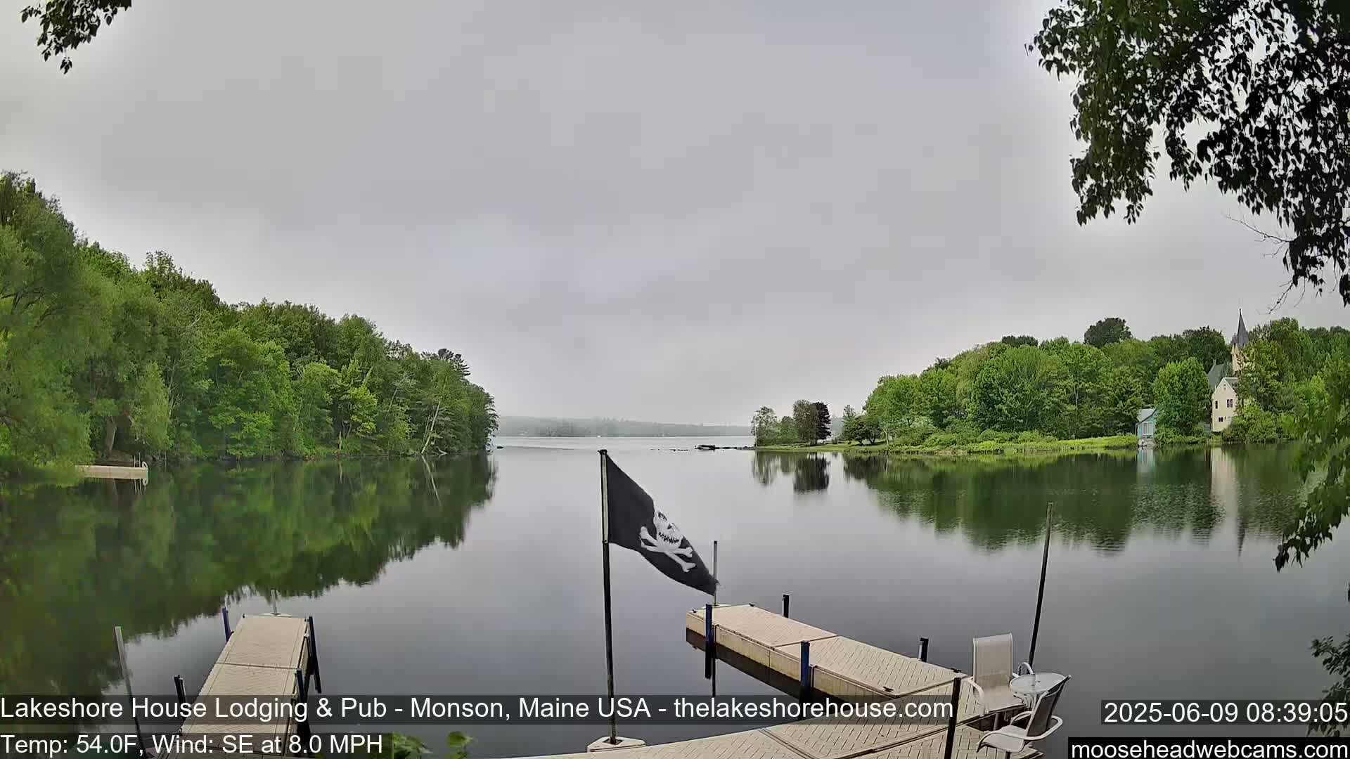 A calm lake reflecting the overcast sky and green trees on its shores, with a small dock and a pirate flag visible.