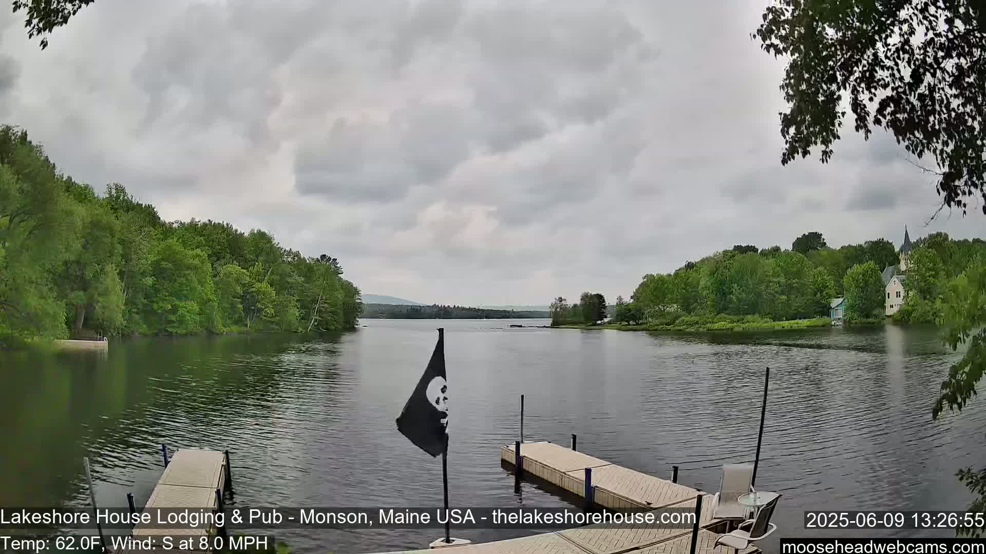 A calm lake is seen from a dock under an overcast sky, with green trees lining the shore and a small flag on the dock.