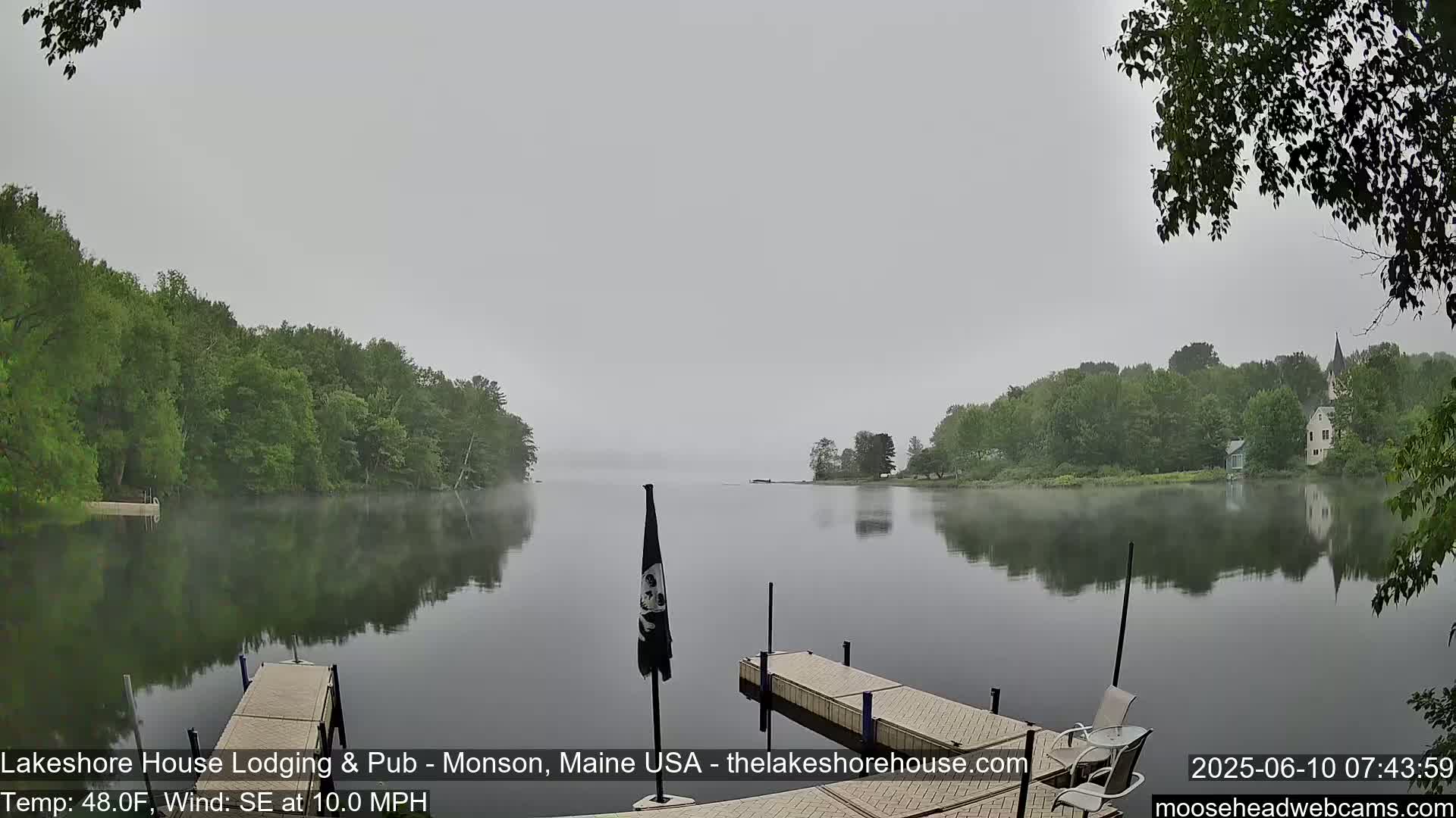 A calm lake shrouded in fog is reflected in the still water, with green trees lining the shore and a wooden dock in the foreground.