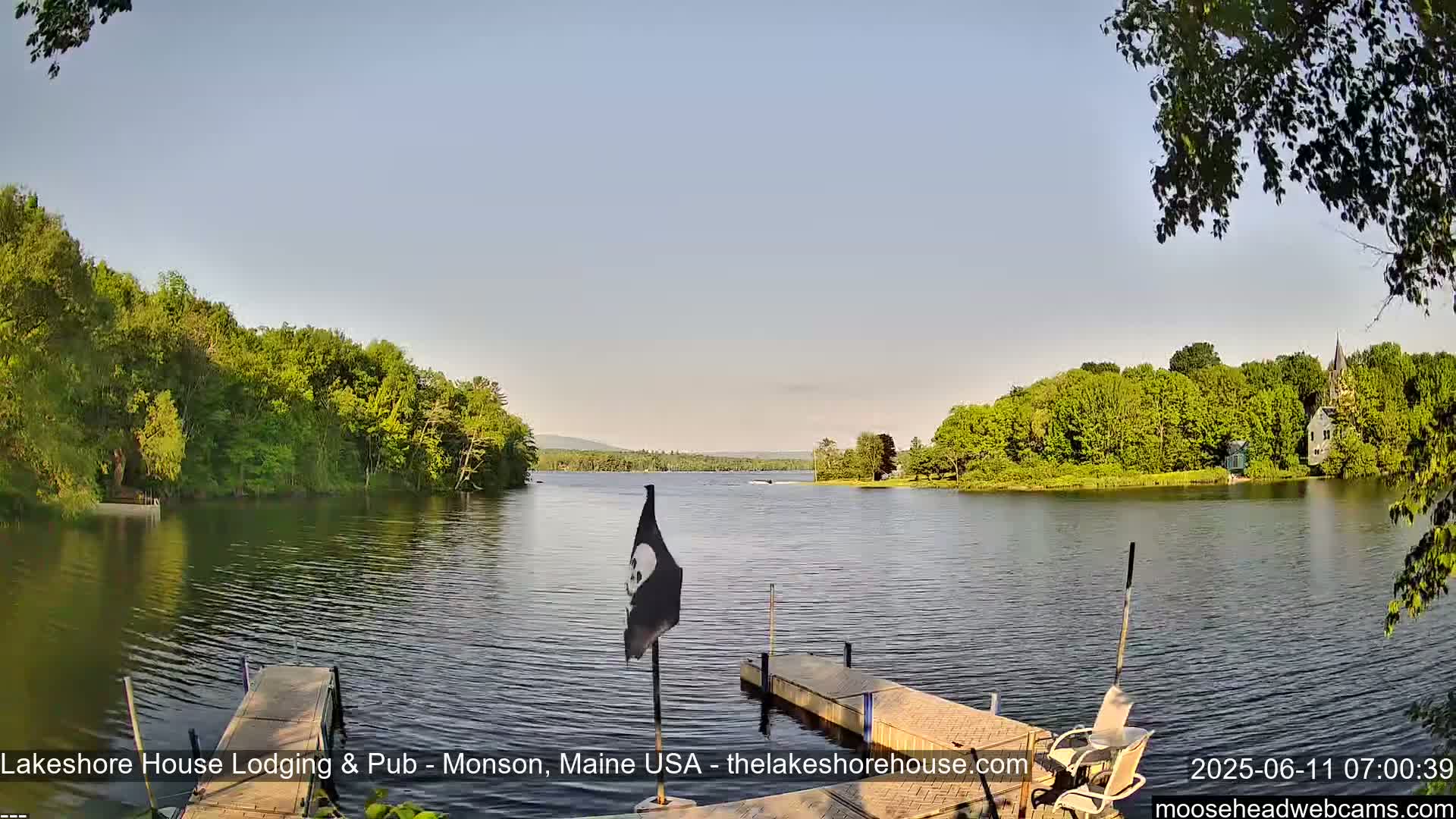A calm lake is surrounded by lush green trees under a clear, light-blue sky, with a small dock and a flag in the foreground.