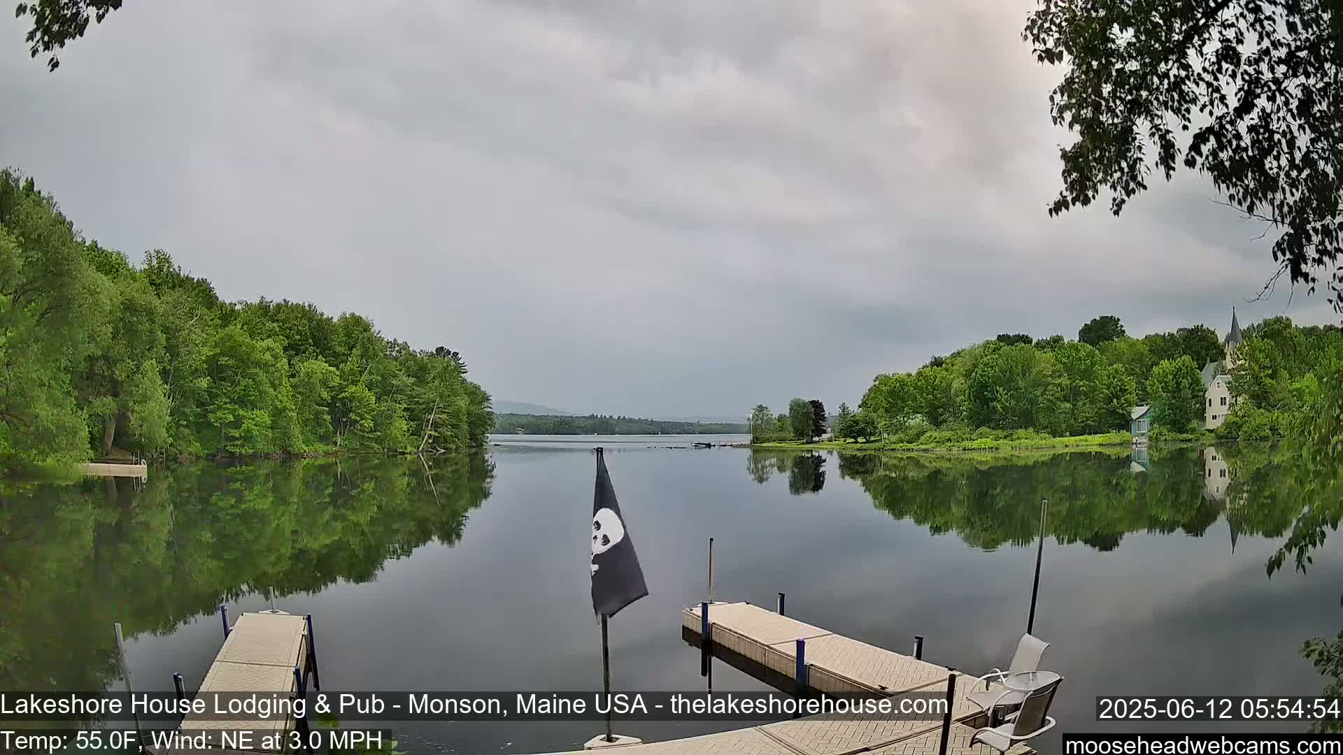 A calm lake reflects a tree-lined shore and overcast sky, with a dock and a black flag in the foreground.