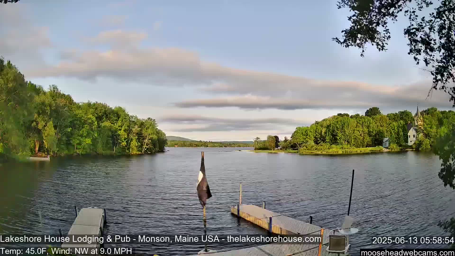 A calm lake, under a partly cloudy sky, is seen from a dock with a flagpole in the foreground;  trees line the shores.