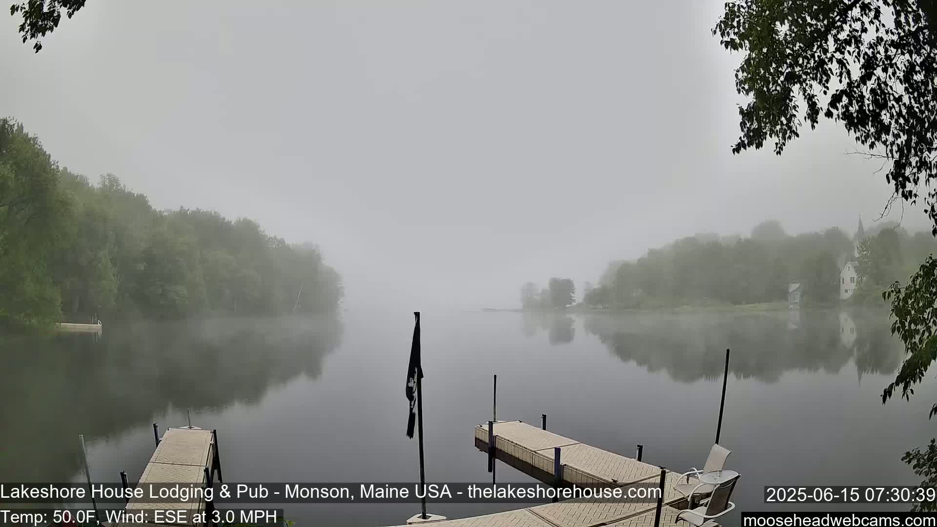 A foggy morning scene shows a calm lake reflecting trees and buildings on the shore, with two docks in the foreground.