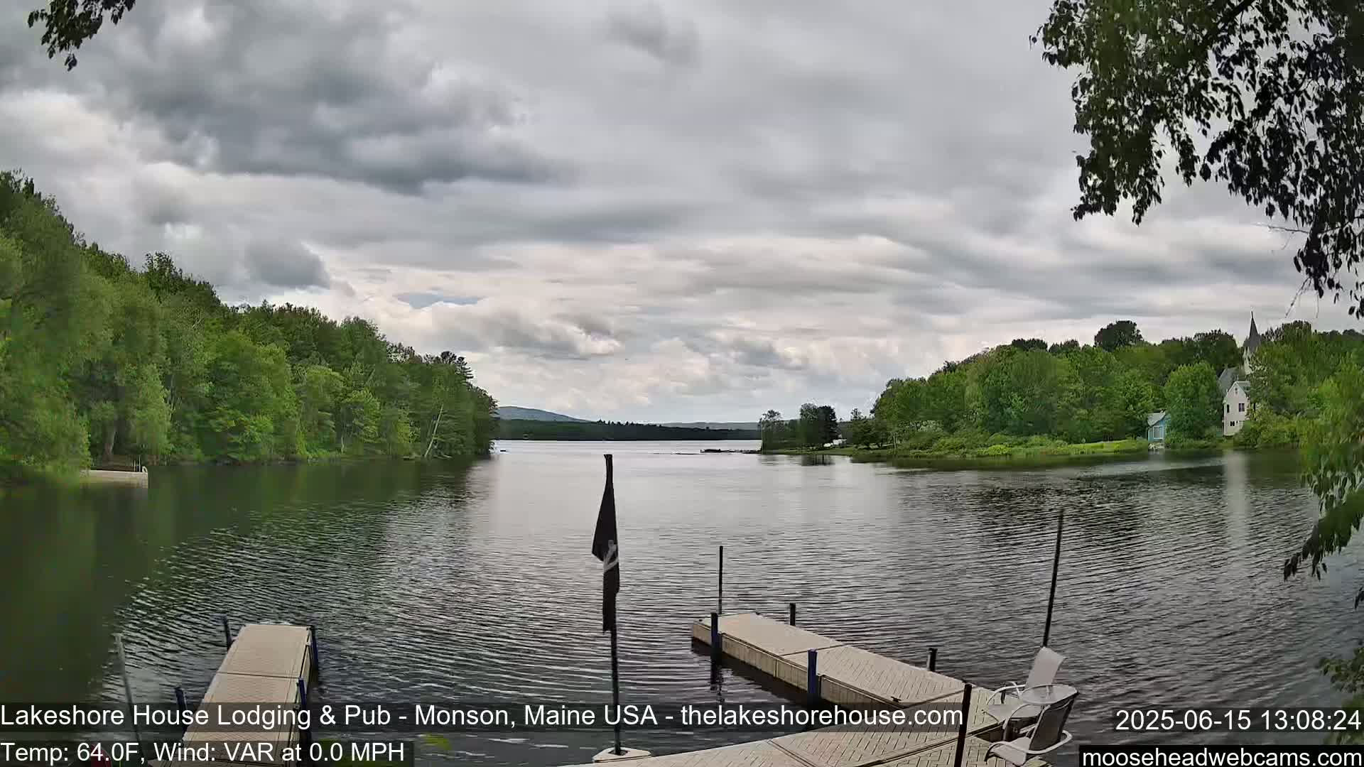 A calm lake under a cloudy sky is viewed from a dock, with lush green trees lining the shoreline and distant mountains visible.