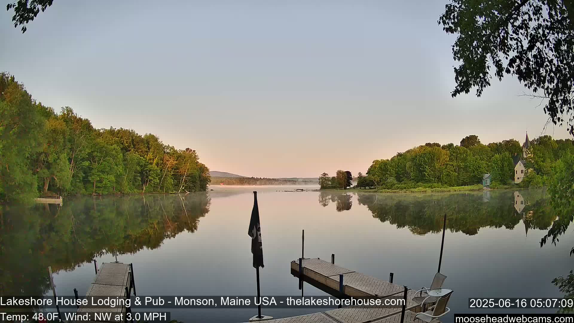 A calm lake at dawn with a light fog, reflecting the trees and buildings on its banks, is viewed from a wooden dock.