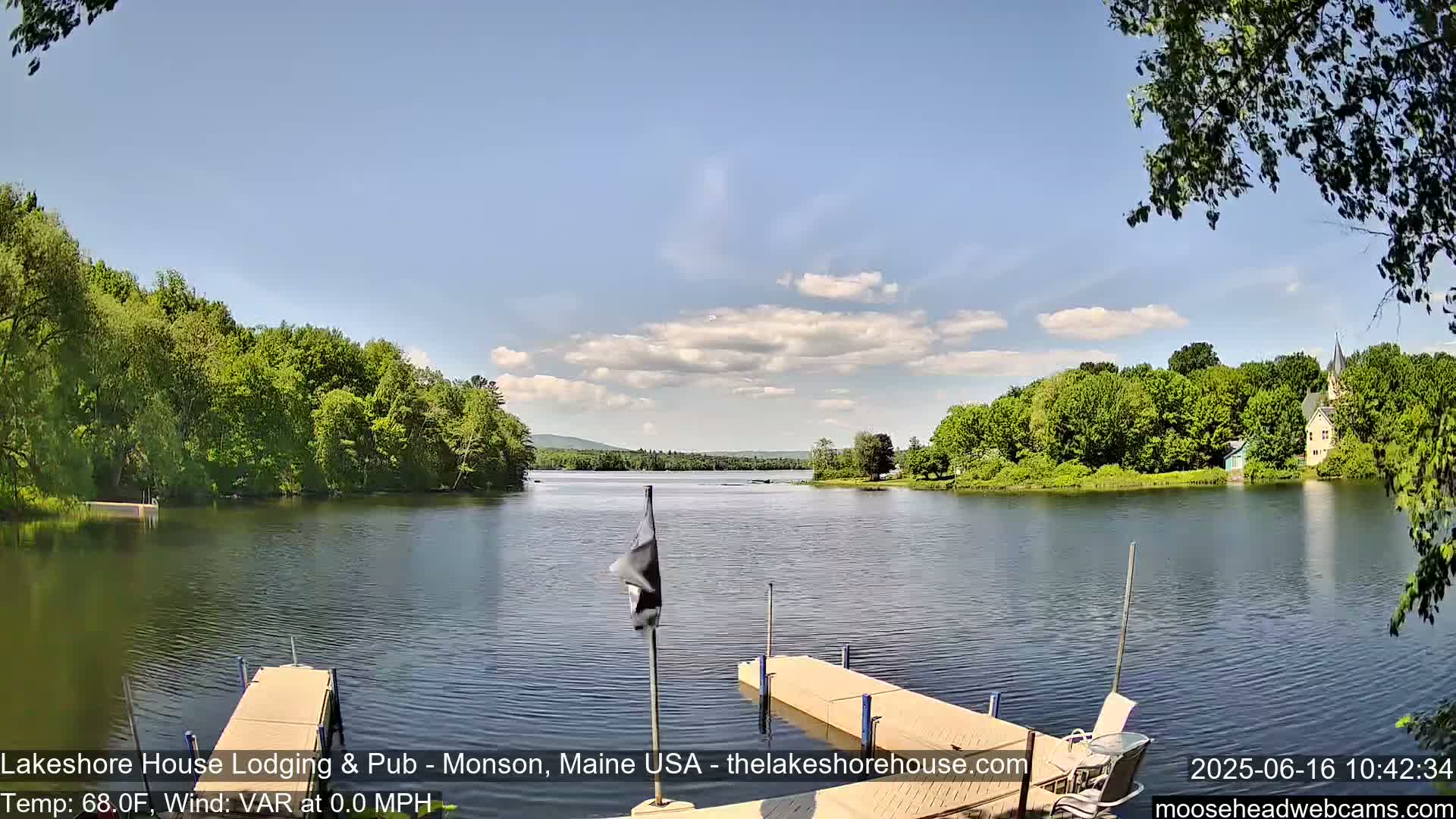 A calm lake is seen from a dock on a partly cloudy day, with lush green trees lining the shores and a building visible in the distance.