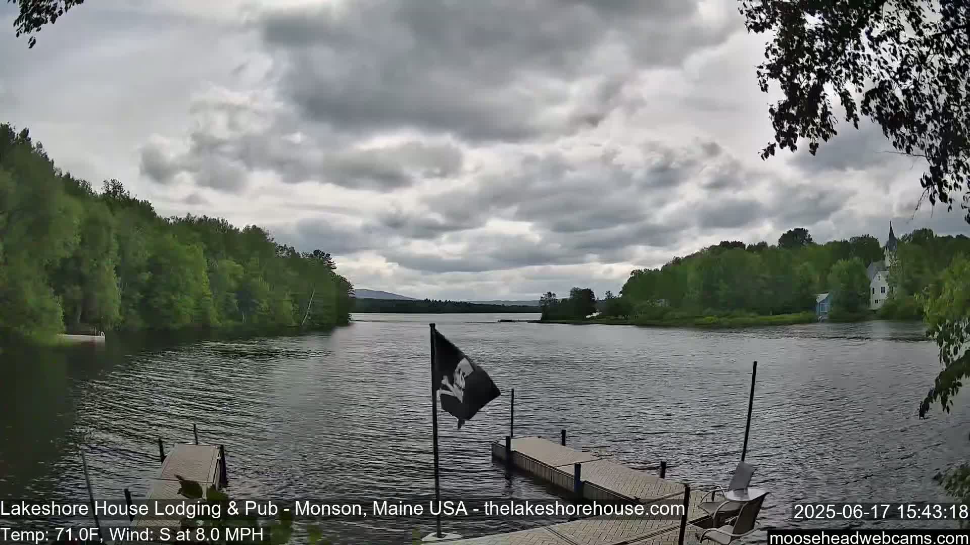 A calm lake is seen under a cloudy sky, with a wooden dock and a pirate flag in the foreground, and lush green trees lining the shoreline.
