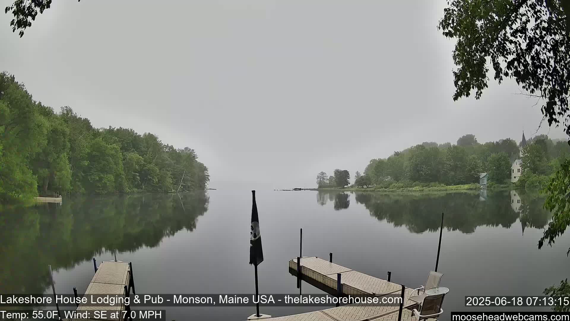 A calm lake, shrouded in fog, is bordered by lush green trees, with a wooden dock extending into the still water.