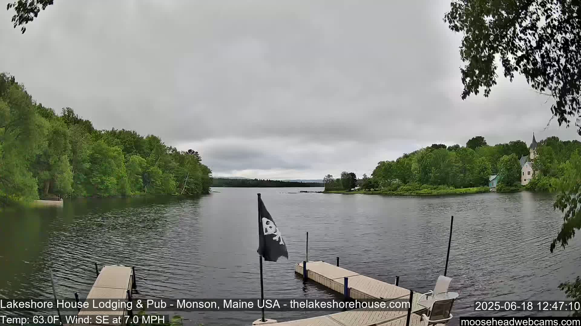 A calm lake under an overcast sky is seen from a dock, with lush green trees lining the shore and a building visible in the distance.