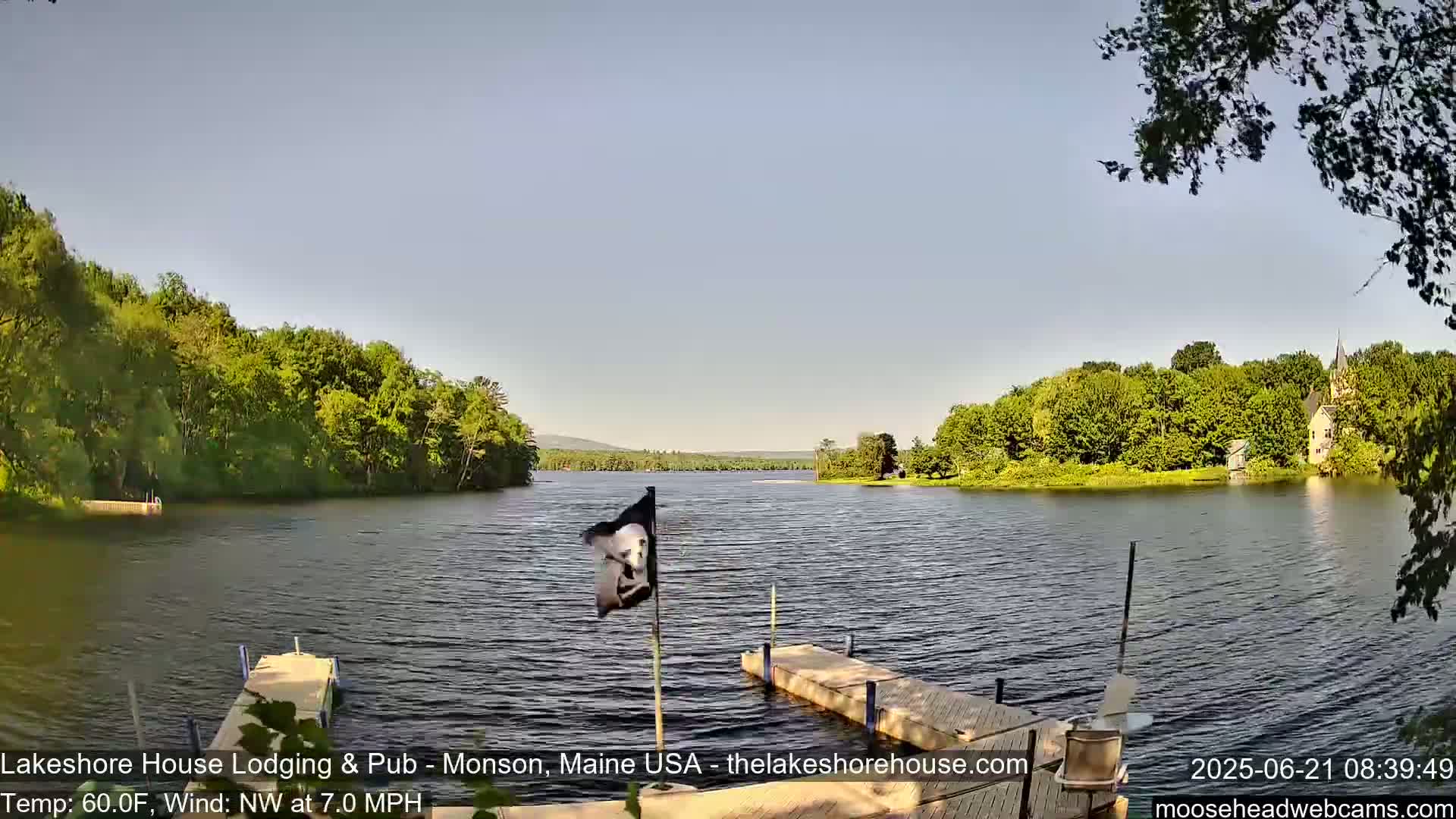 A calm lake with green wooded shores and a small dock is visible under a clear sky, with a gentle northwest wind.