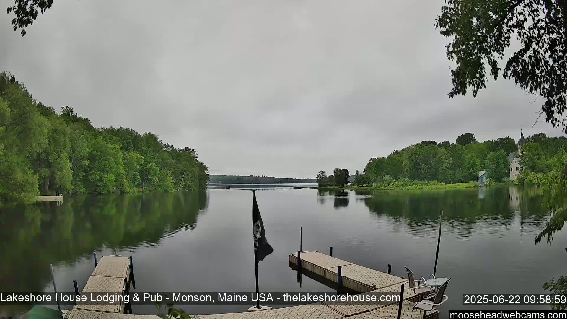 A calm lake is seen under an overcast sky, bordered by lush green trees and featuring a dock in the foreground.