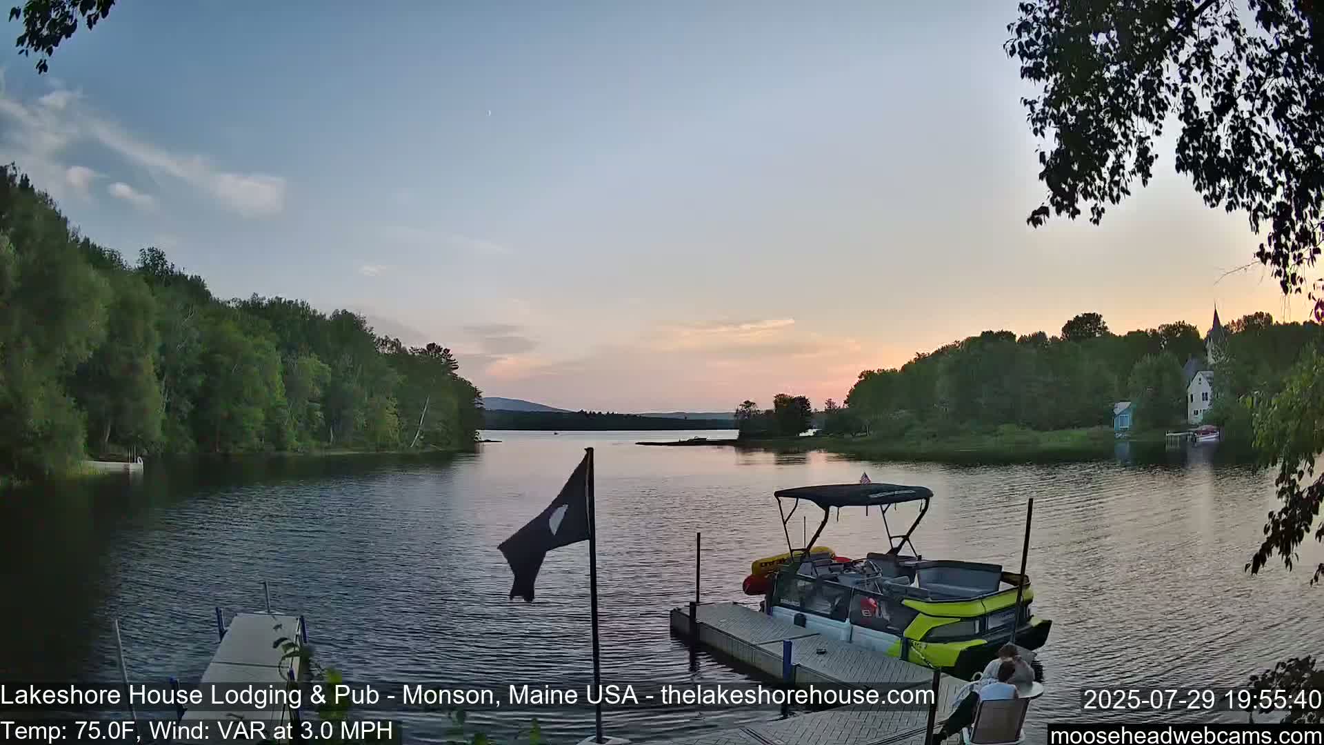 A pontoon boat is docked on a calm lake surrounded by lush green trees under a bright, sunny sky.