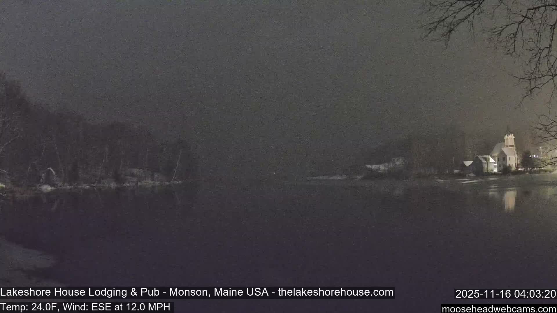 A hazy night scene reveals a calm body of water reflecting the lights of a white church with a steeple and other buildings on the distant bank, flanked by bare trees and patches of snow on the opposing shore.