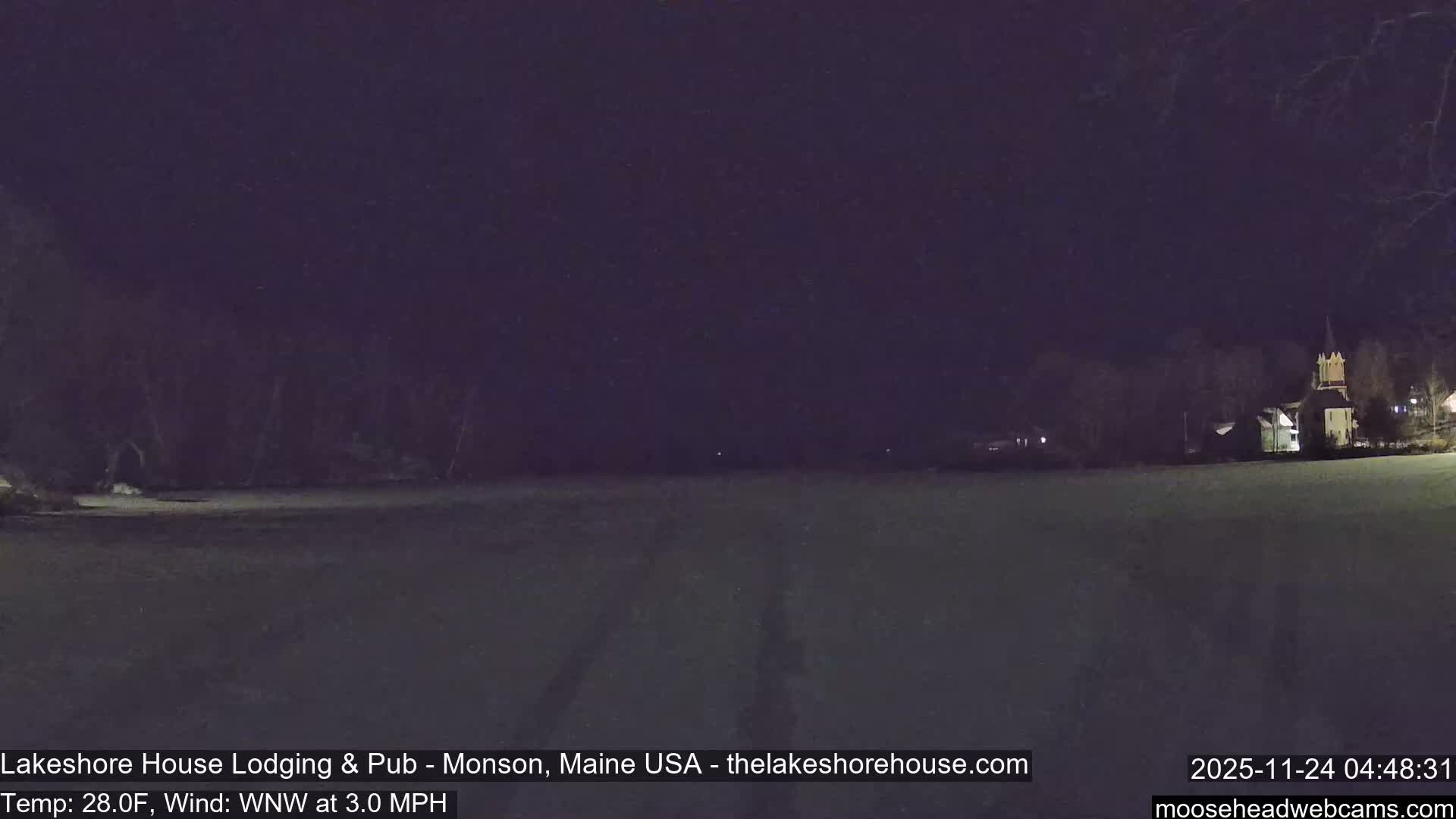 On a cold, clear night, a snowy or icy expanse stretches into the distance, bordered by dark trees, with a brightly lit church and other buildings nestled on the right shore.