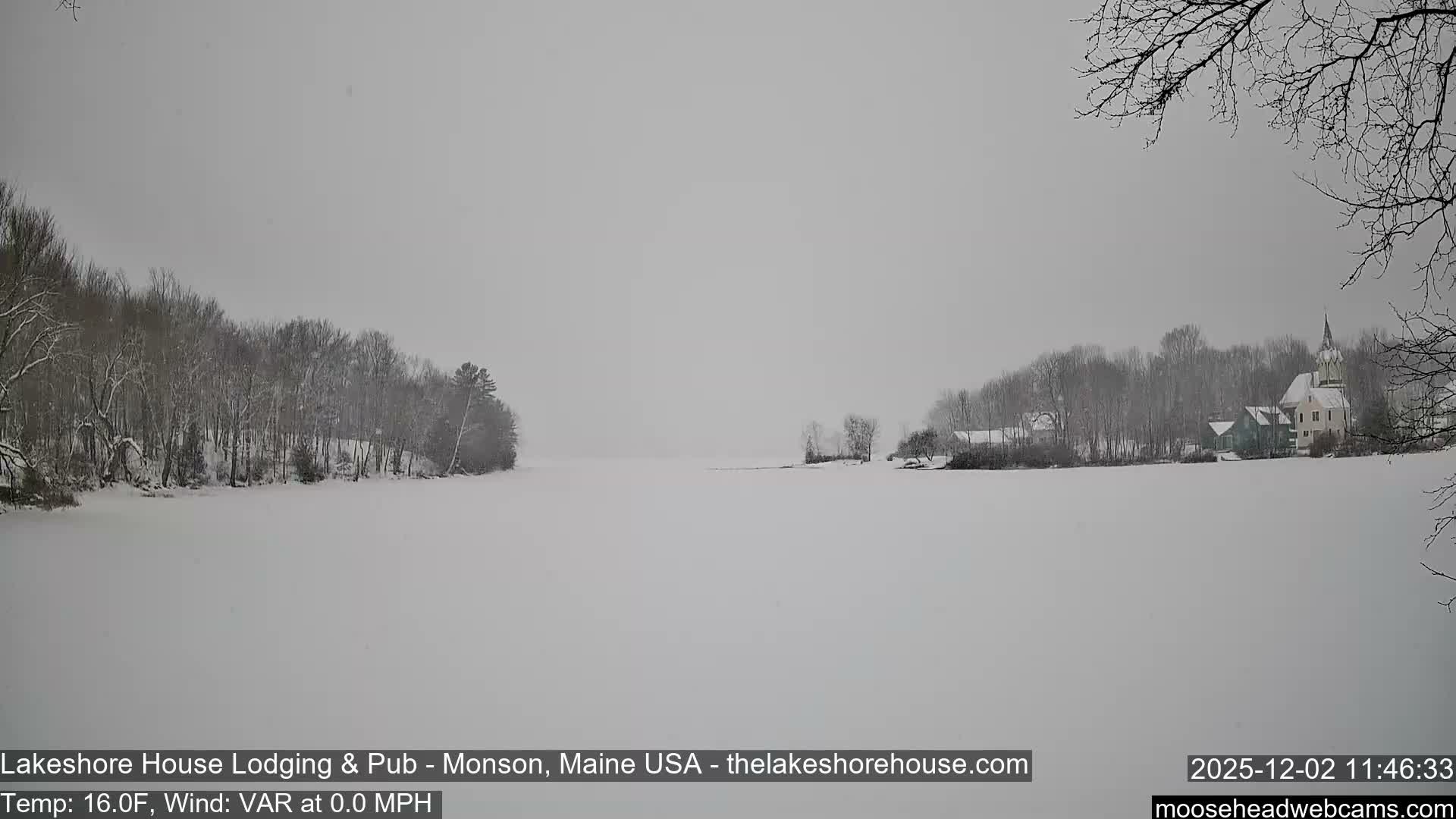 A snow-covered landscape on an overcast day features a vast frozen lake or field in the foreground, bordered by bare trees on the left and a small collection of buildings including a prominent church steeple on the right, all dusted with fresh snow under a gray sky.