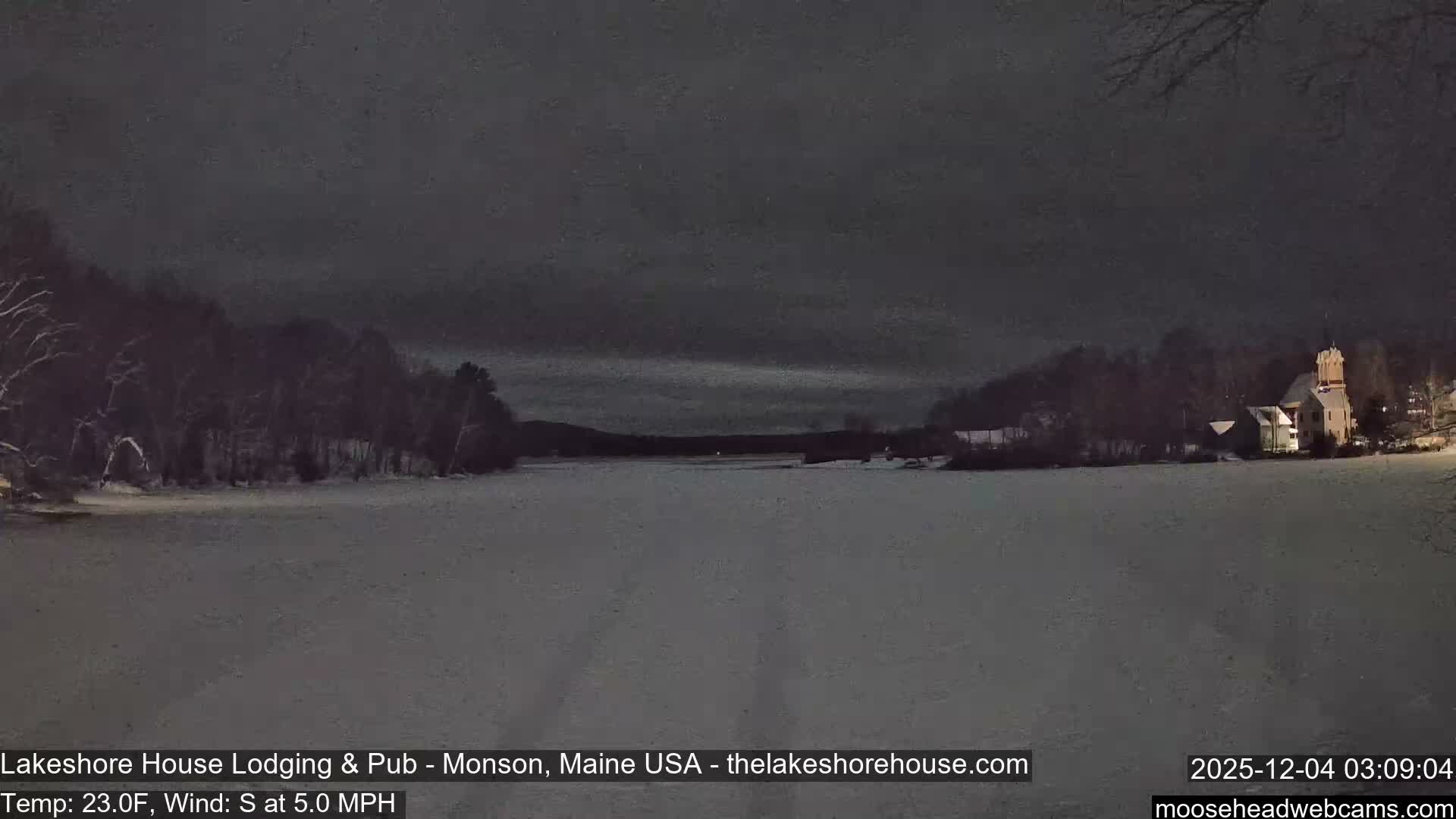 A snowy winter landscape at night features a frozen expanse (likely a lake) flanked by snow-dusted trees, with a distinctive illuminated building on the right under a dark, overcast sky.