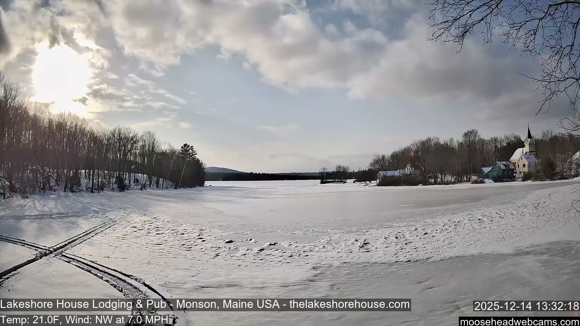 A snowy winter landscape at night features a frozen expanse (likely a lake) flanked by snow-dusted trees, with a distinctive illuminated building on the right under a dark, overcast sky.