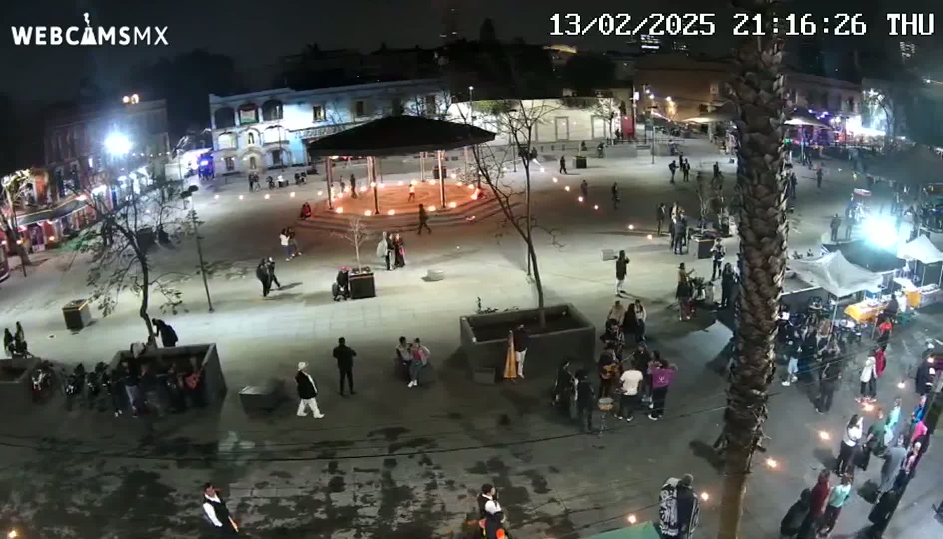 A nighttime aerial view of a town square with many people gathered around a gazebo, small fires, and various groups of musicians.