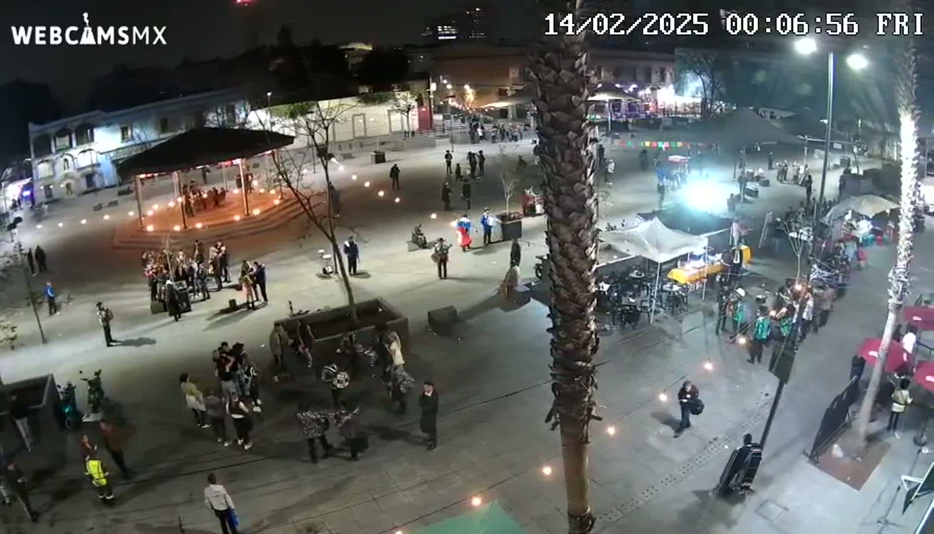 A nighttime aerial view of a town square with numerous people gathered around various groups, including musicians and vendors, under illuminated lights.