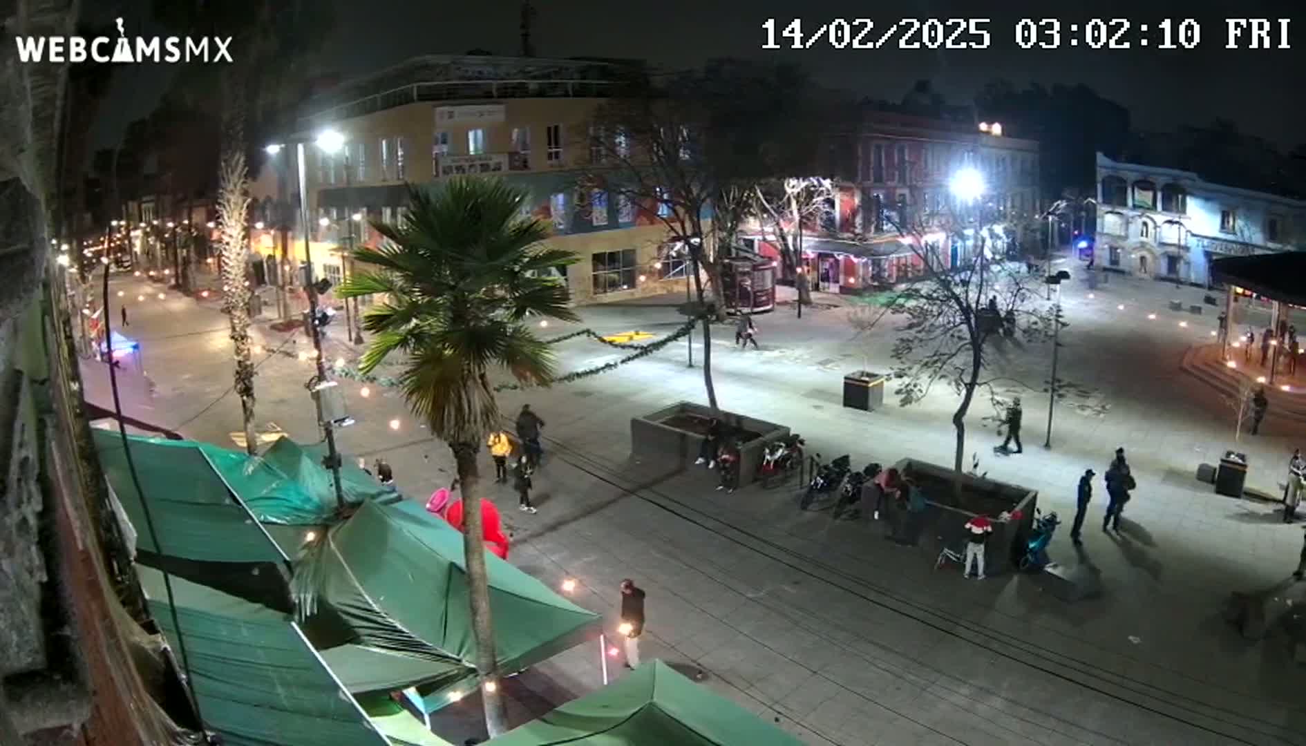 A nighttime, illuminated town square with several people scattered throughout, some under temporary tents, and palm trees lining a street.