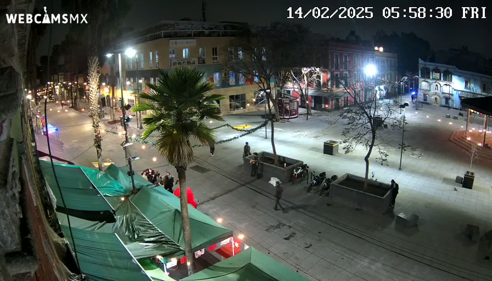 A nighttime view of a plaza with several buildings, a few people, some parked motorcycles, market stalls with green canopies, and a palm tree, illuminated by streetlights.
