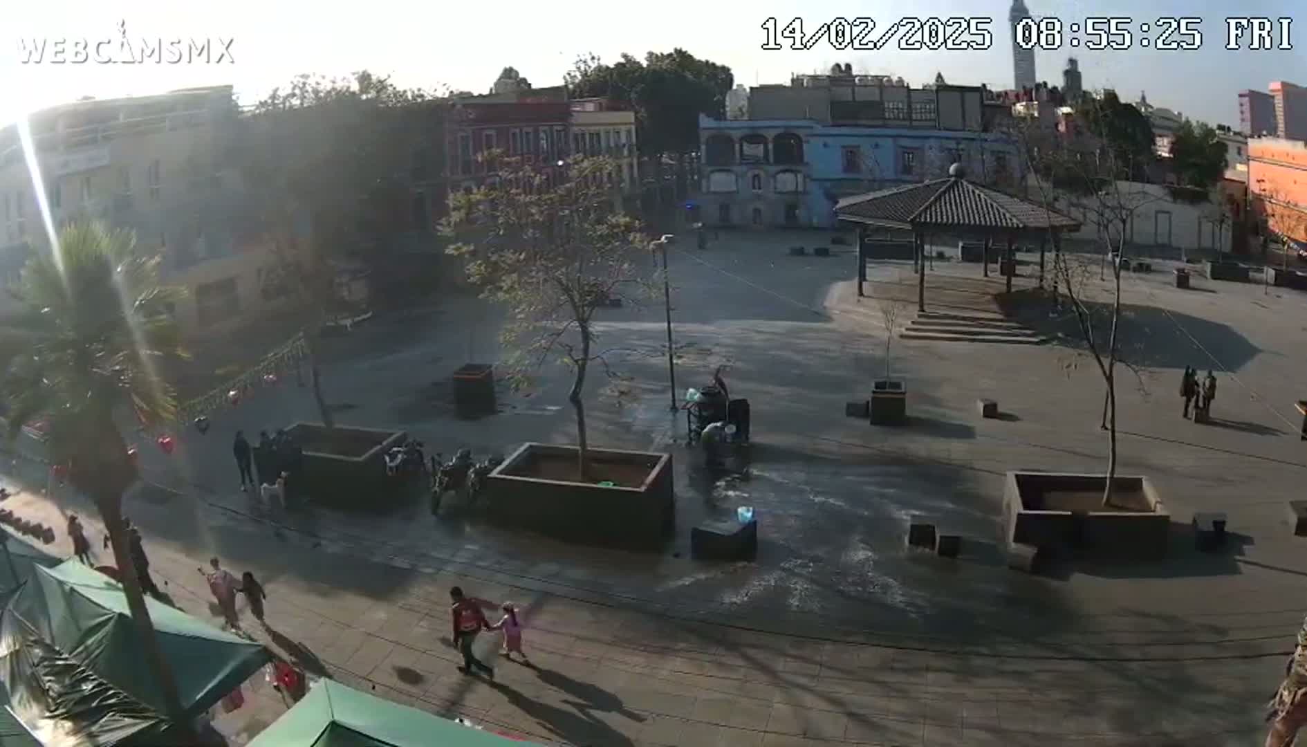 A sunny day reveals a plaza with a gazebo, several people walking around, and buildings in the background.