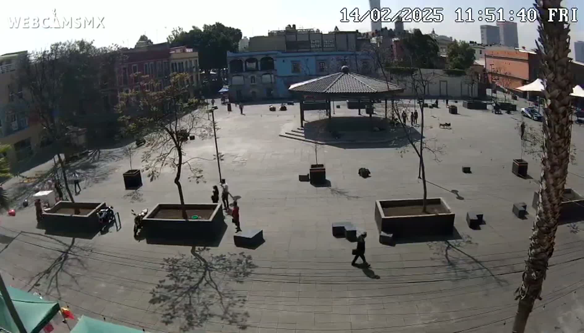 A mostly empty plaza with a gazebo in the center, surrounded by low buildings on a sunny day.