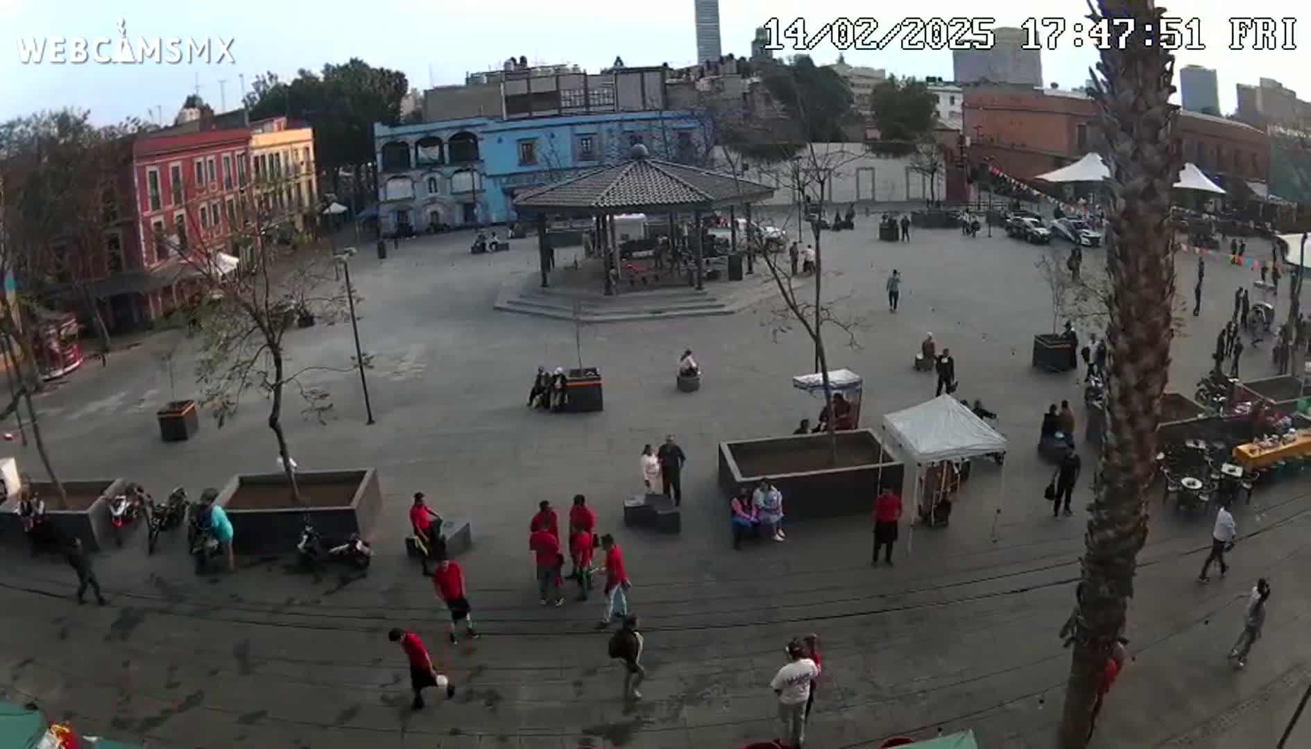 An overcast day shows a town square with several people walking around, a gazebo in the center, and various small stalls and vendors set up around the perimeter.