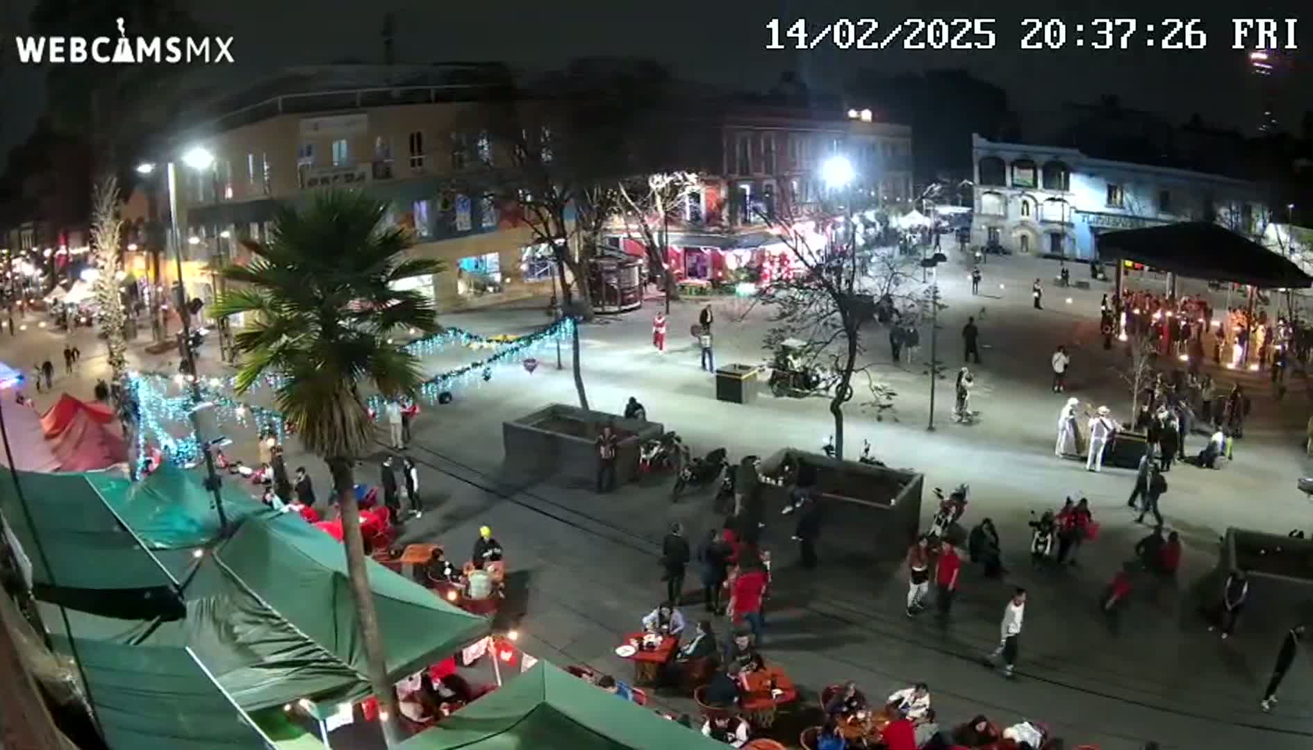 A nighttime aerial view of a city square with many people gathered, some sitting at outdoor tables, under illuminated buildings and festive lights.
