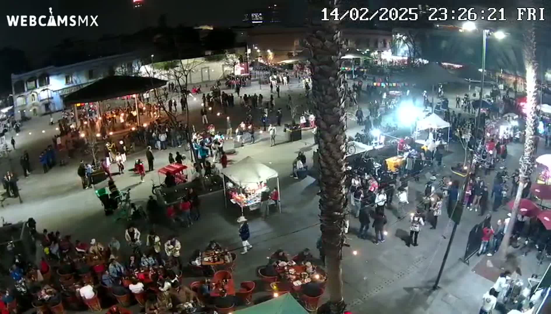 A nighttime, aerial view of a crowded plaza with numerous people gathered around various stalls, vendors, and seating areas under ambient lighting.