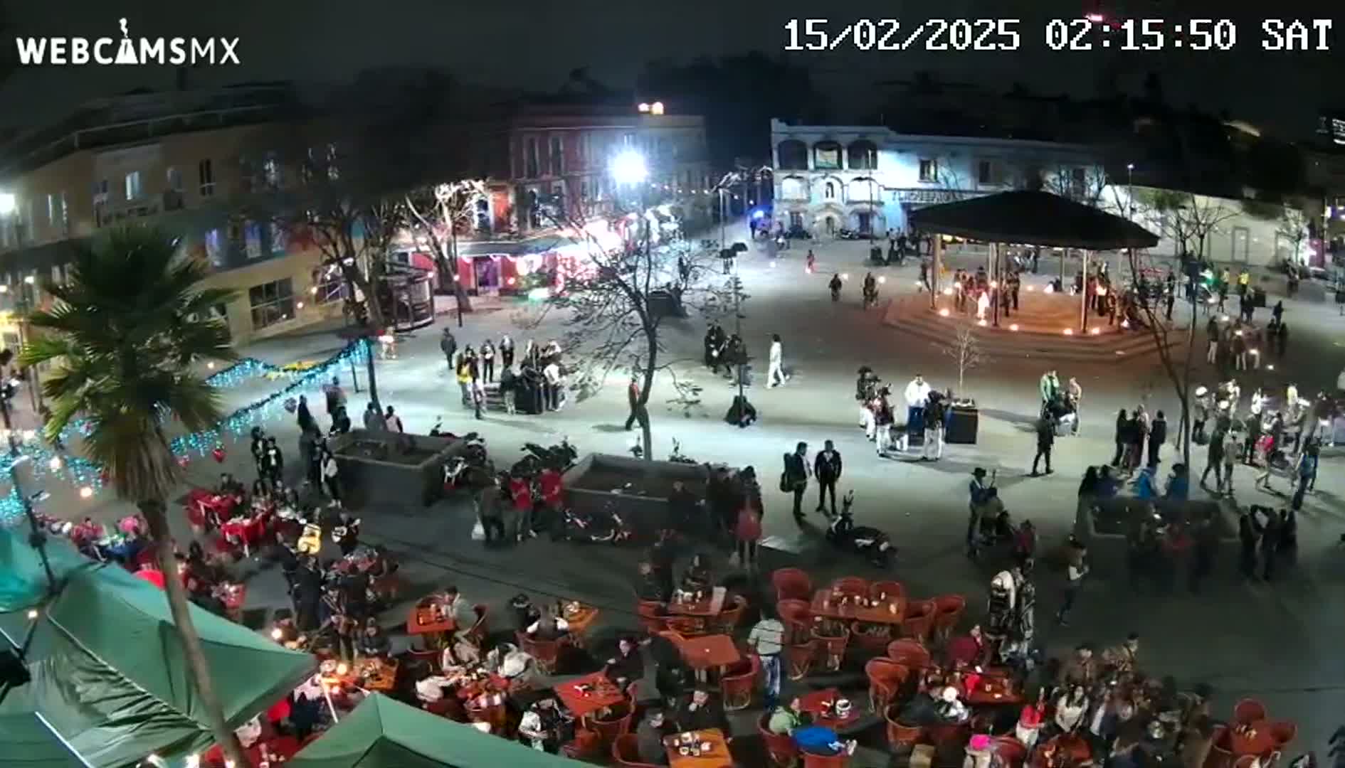 A nighttime aerial view of a town square with many people gathered around outdoor seating areas and a gazebo, under clear skies.