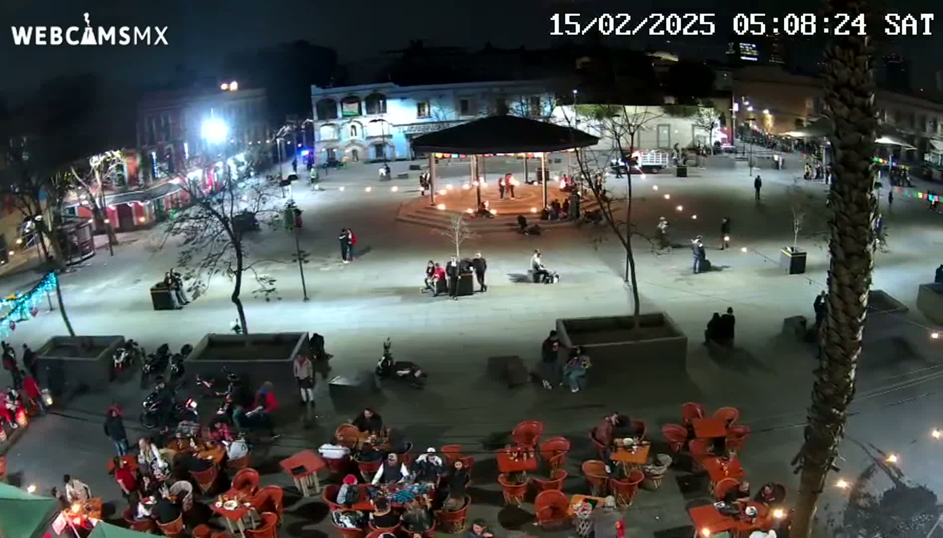 A nighttime aerial view of a town square with people gathered around tables and a gazebo, under clear skies.