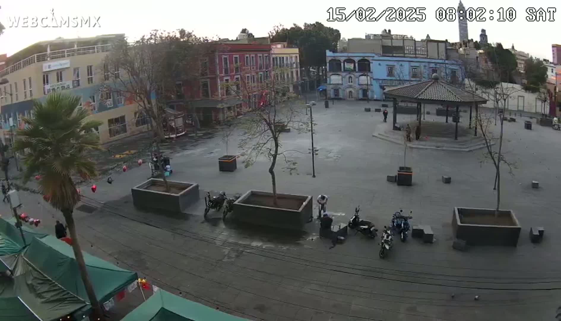 An overcast day reveals a town square with several parked motorcycles, a gazebo, and low buildings surrounding the paved area, with a palm tree and some market stalls visible on the street.