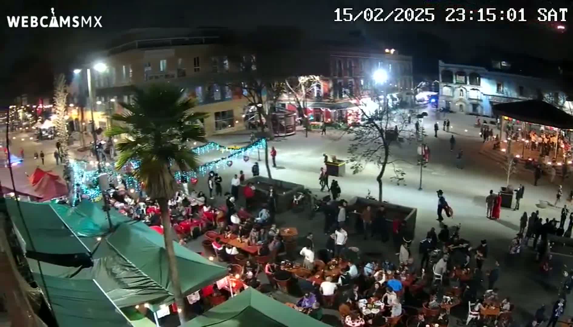 A nighttime aerial view of a town square filled with people at outdoor restaurants and stalls under clear skies.